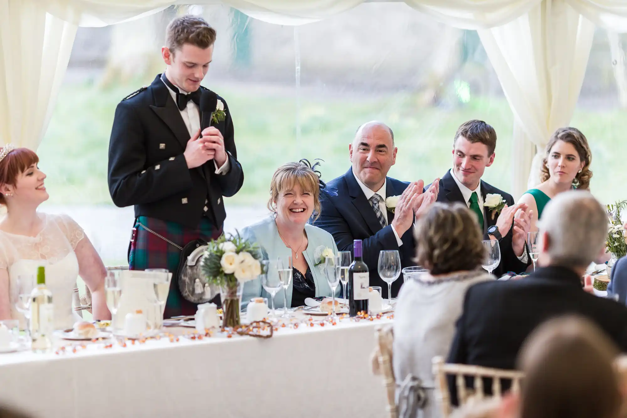 A young man in a kilt stands giving a speech at a formal event, likely a wedding. He holds a microphone and is smiling. Seated guests dressed formally, some applauding and smiling, are at a decorated long table with flowers, glasses, and bottles.