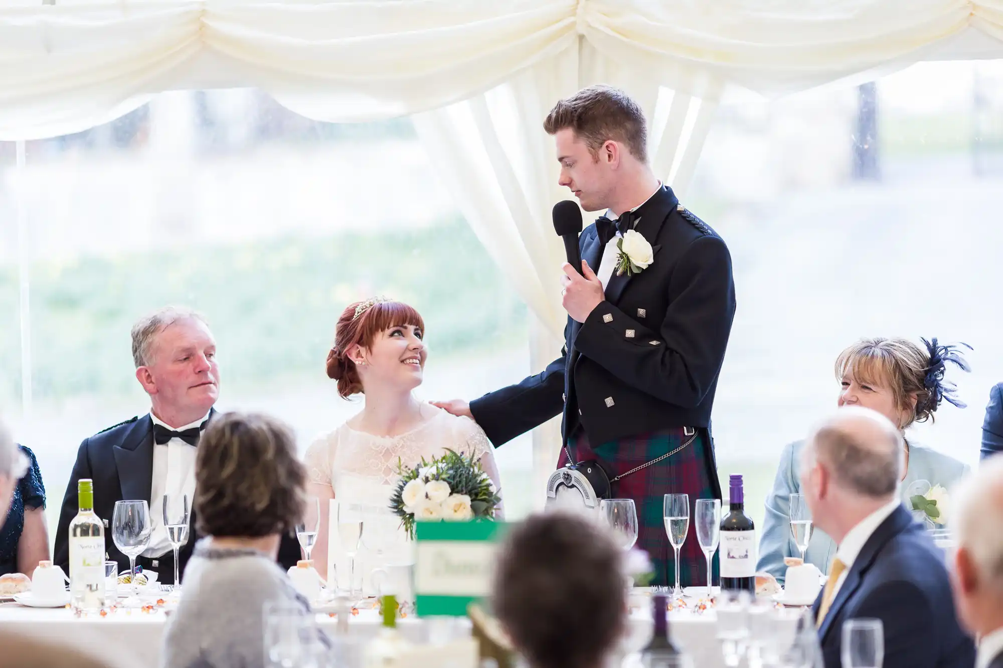 A groom in a black jacket and kilt stands holding a microphone, smiling at a seated bride in a white dress holding a bouquet. They are at a wedding reception table with guests, drinks, and decorations visible under a white marquee.