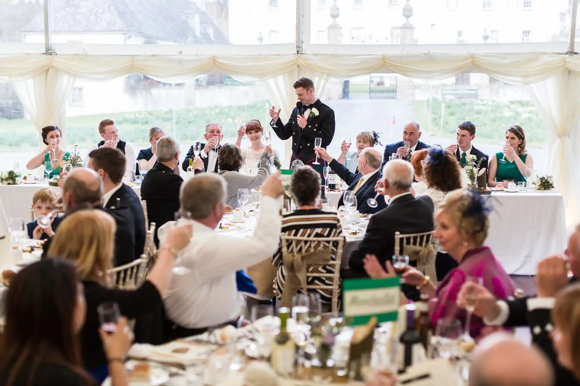 A man in a black suit stands and gives a toast at a long, formal wedding reception table. Guests seated and facing him raise glasses in celebration. The room has white draped curtains, and the atmosphere is lively and festive.