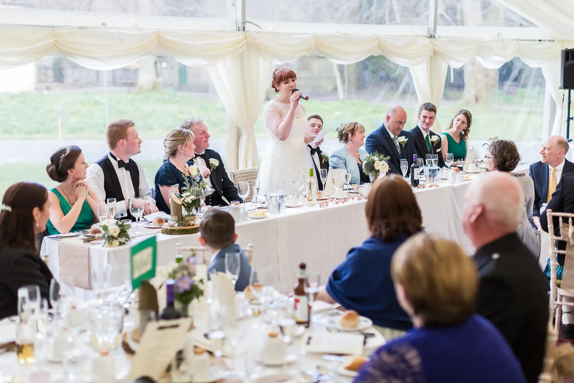 A bride stands at a long table, speaking into a microphone at a wedding reception. Guests in formal attire sit around the table, decorated with flowers, glasses, and plates. A bright white marquee with large windows is visible in the background.