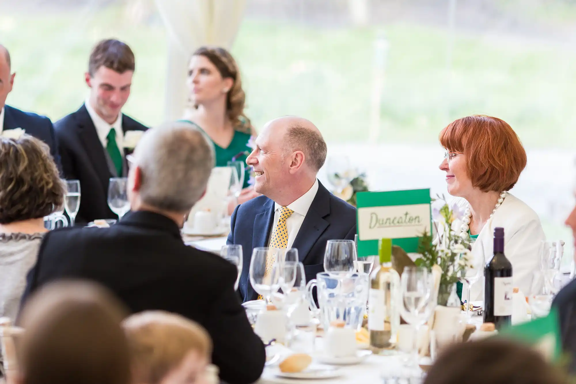 A group of well-dressed people sit around a formal table at an event, smiling and talking. The table is set with glasses, plates, wine, and a green card labelled "Duncaton." The background is bright and softly blurred.