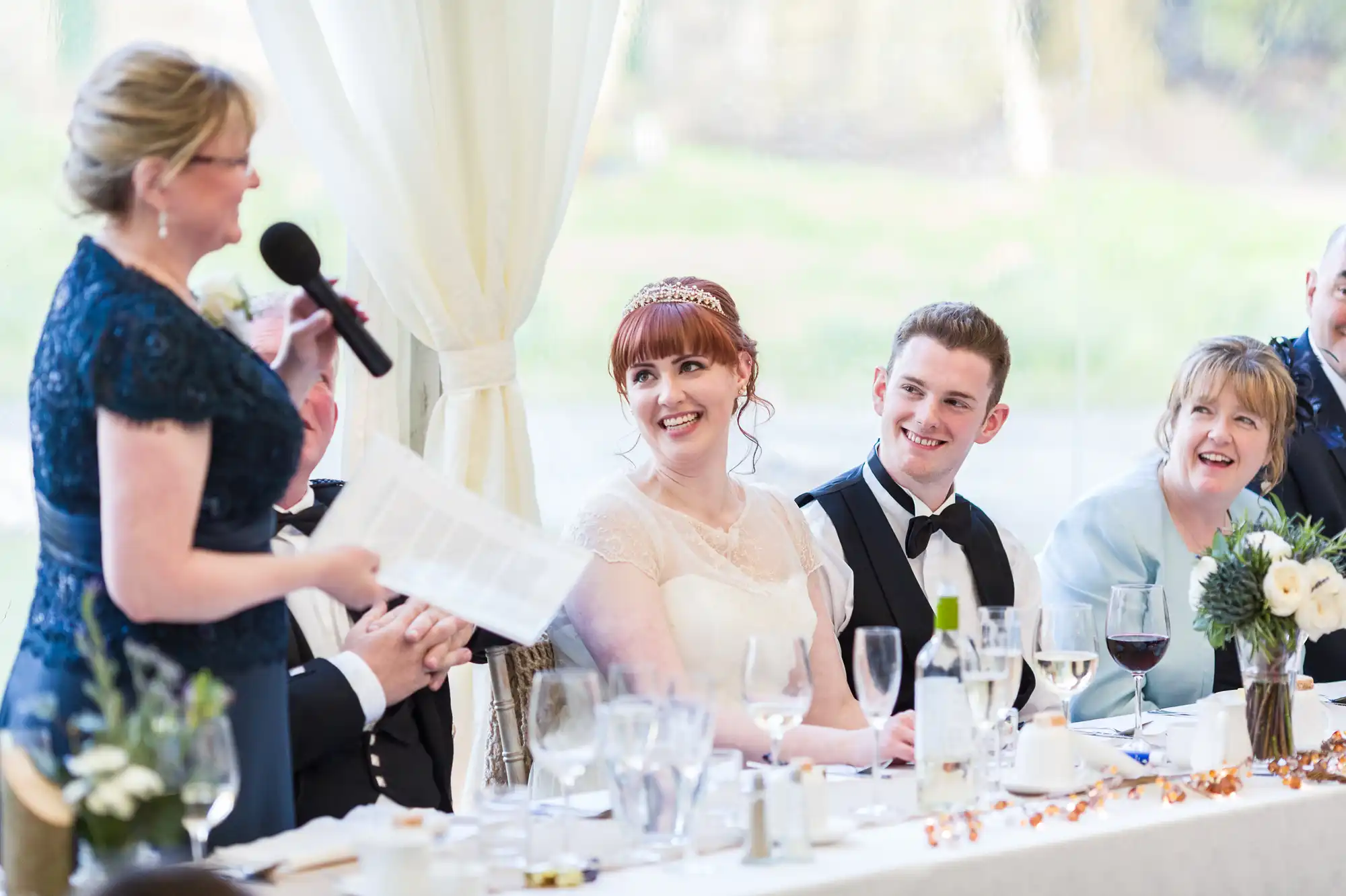 A woman in a blue dress speaks into a microphone at a wedding reception table. The bride and groom, seated among guests, smile warmly at her. The table is set with glasses, plates, floral arrangements, and festive decorations.