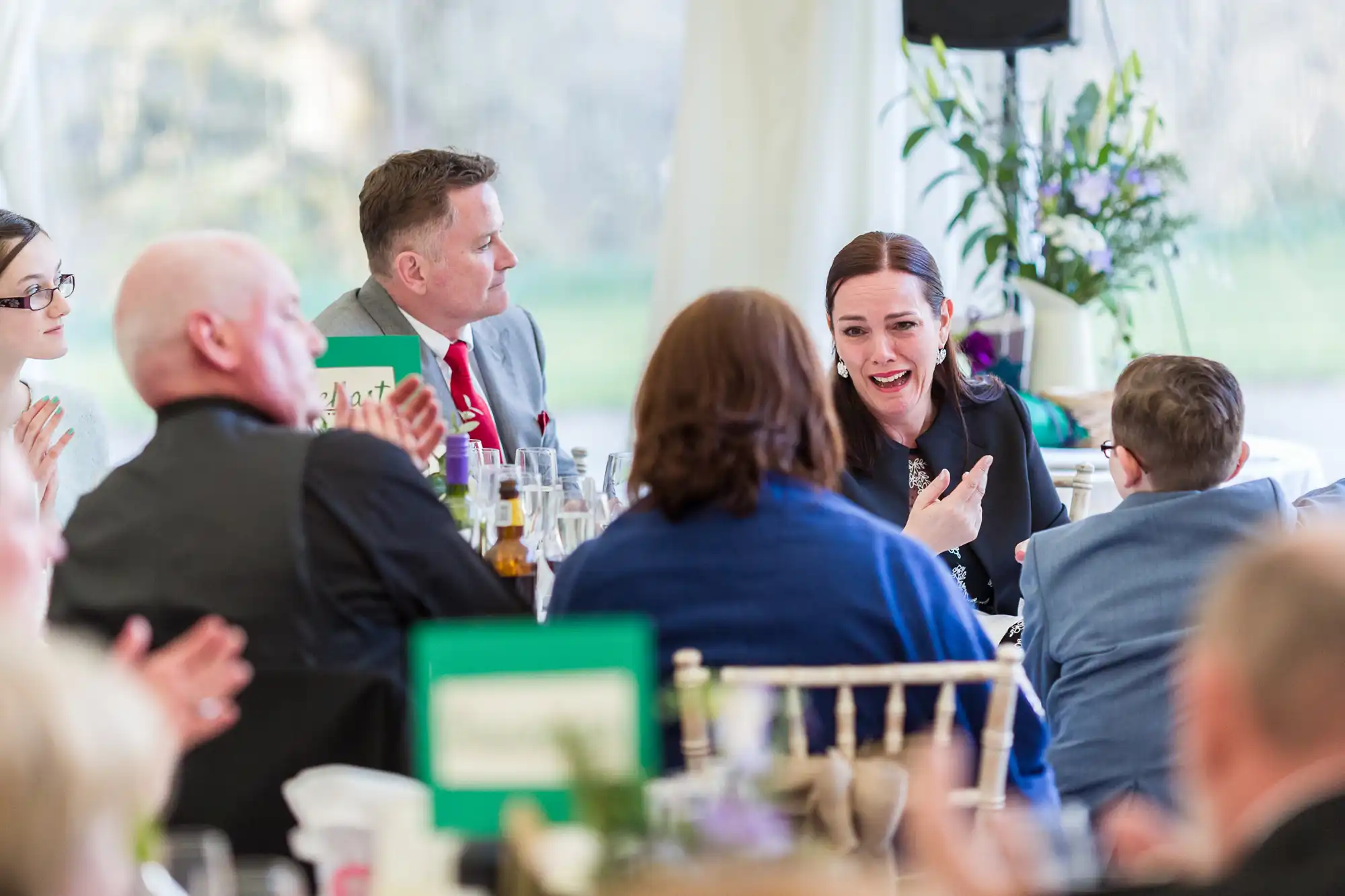 A group of people sit round a table at a formal event, dressed in suits and smart clothing. A woman in black is laughing and gesturing, while others are smiling, clapping, and listening. Bottles and glasses are on the table, and flowers decorate the background.