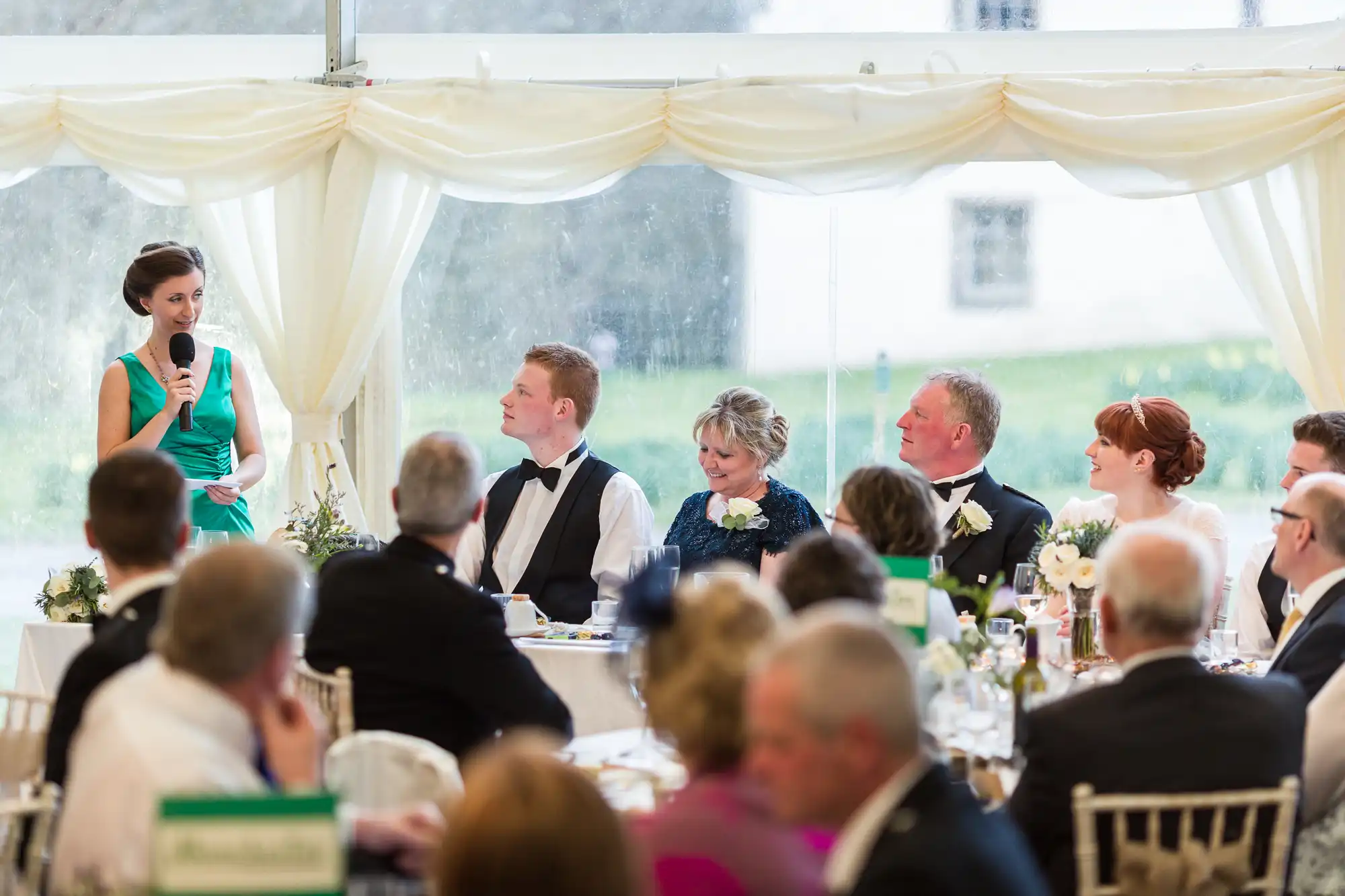 A woman in a teal dress speaks into a microphone at a wedding reception, addressing a long table where the bride, groom, and their families, all dressed formally, sit and listen. Guests are seated in the foreground, and white drapery decorates the marquee venue.