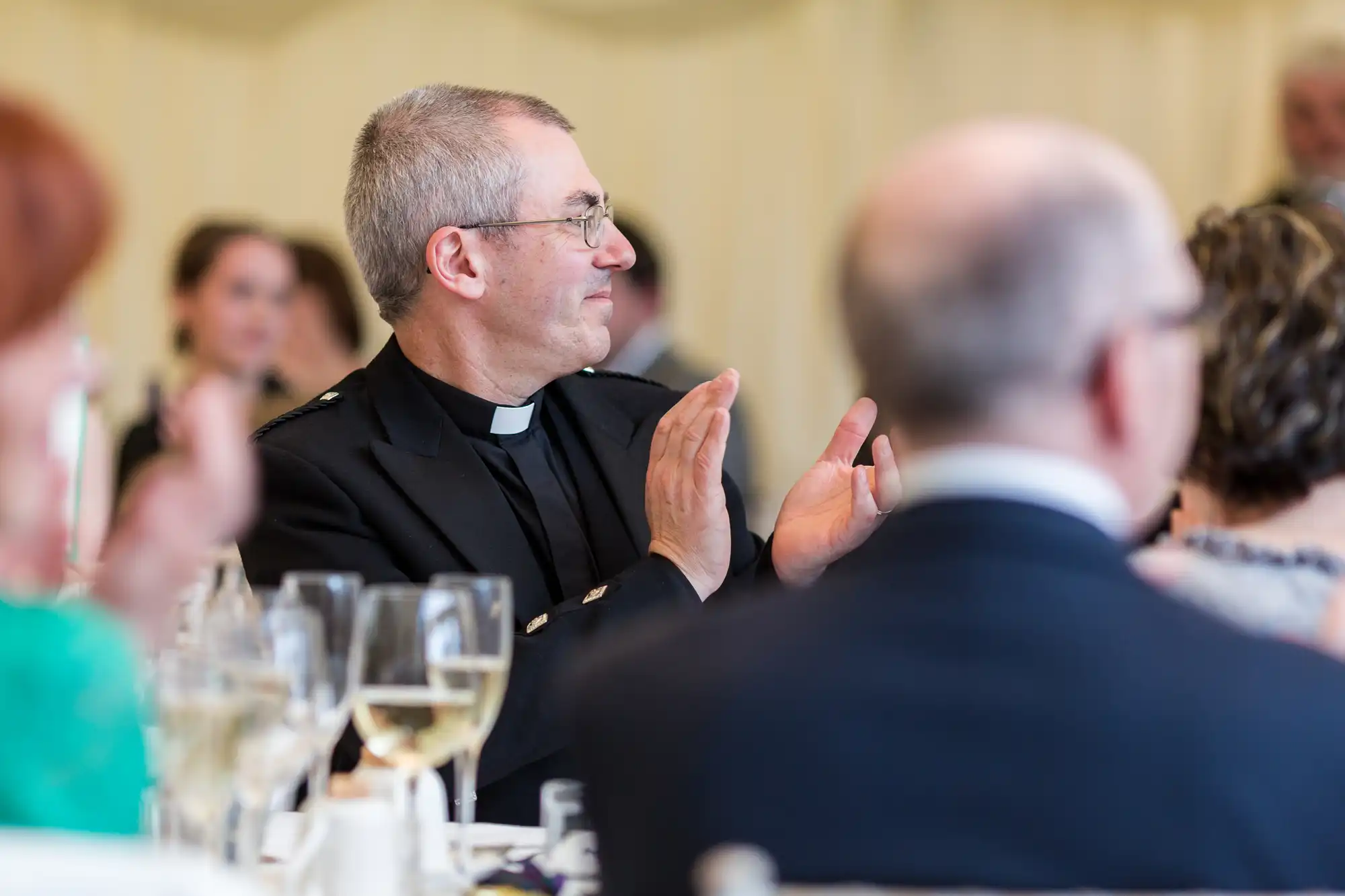 A man in a clerical collar and glasses is clapping at a formal event, seated at a table with wine glasses and people around him. The background is softly blurred, highlighting the man's smiling profile and capturing an elegant, celebratory atmosphere.