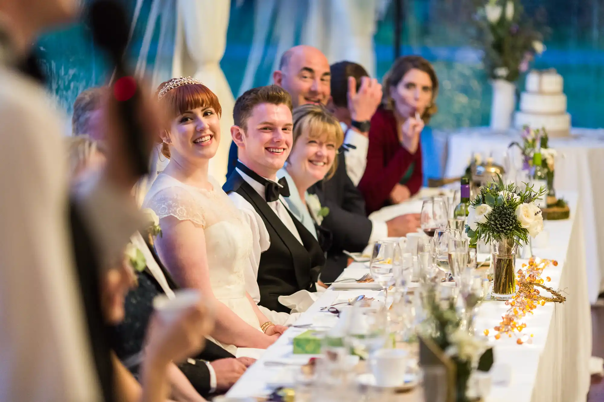 A bride and groom sit smiling at a decorated banquet table with guests during a wedding reception. People beside them are also smiling and looking towards a speaker in the foreground. The table is adorned with flowers, candles, and glassware, with a wedding cake in the background.