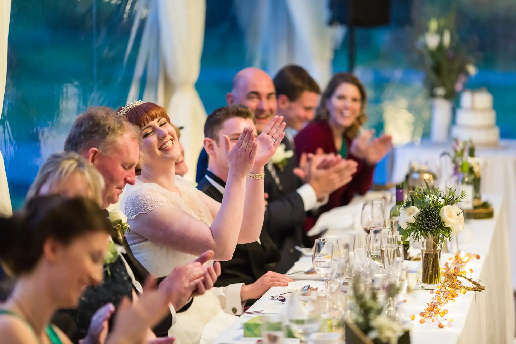 A joyful bride in a white dress sits at a long table, clapping and smiling with guests at a wedding reception. The group, dressed formally, shares laughter and applause. Elegant table settings and floral arrangements decorate the scene inside a bright, marquee venue.