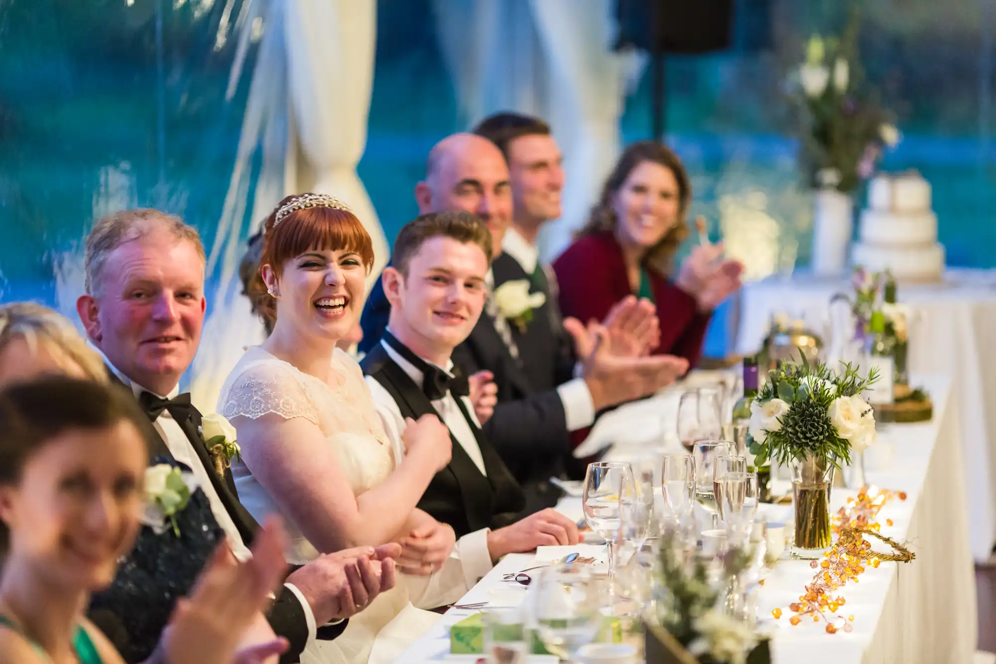 A joyful bride in a white dress sits beside the groom at a decorated banquet table, surrounded by wedding guests in formal attire. People smile, clap, and engage, with flowers, glassware, and a wedding cake visible in the brightly lit background.
