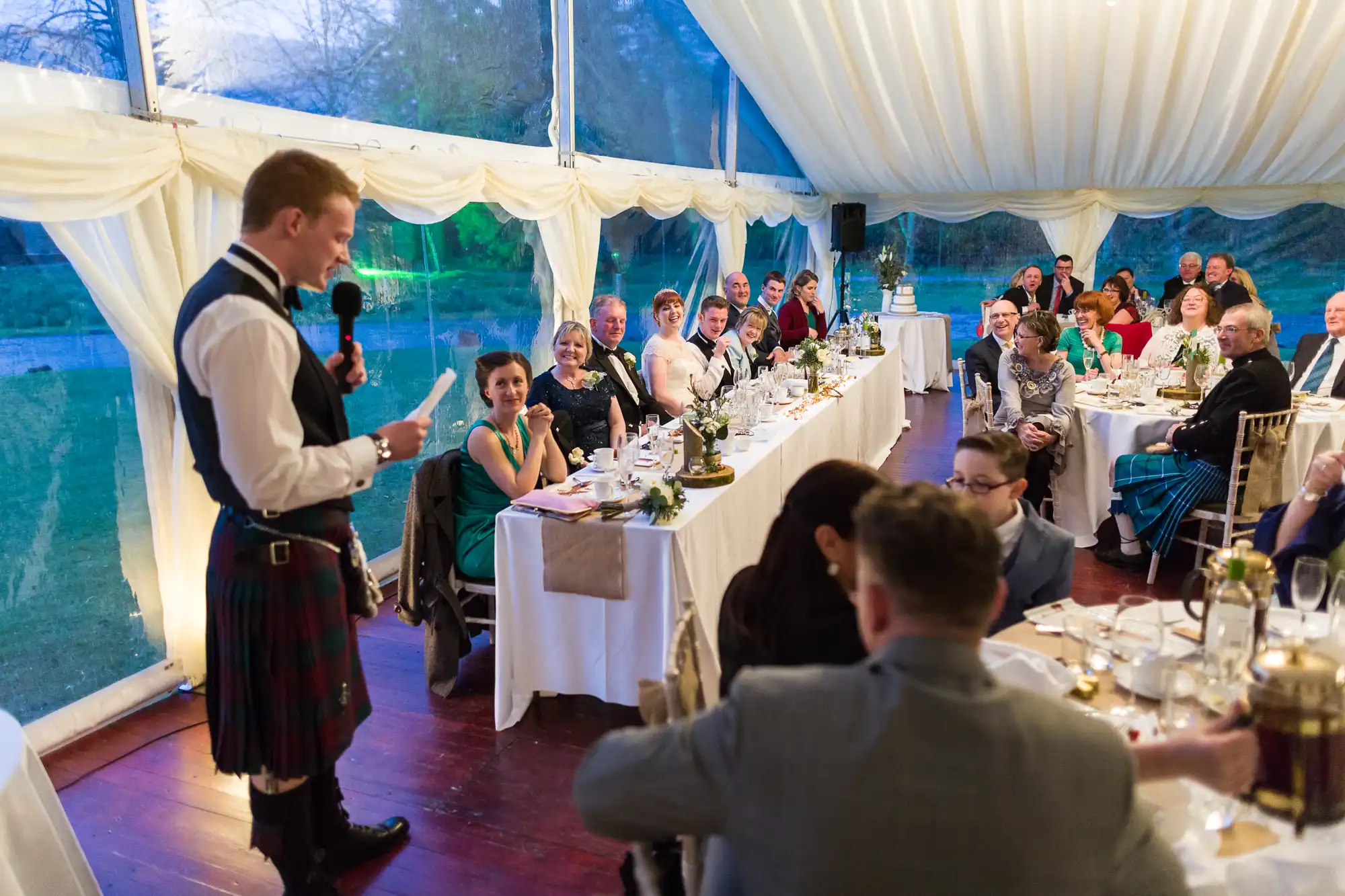 A man in a kilt stands speaking into a microphone at a wedding reception in a marquee. Guests seated at round and rectangular tables listen attentively, smiling. The tent has white drapes, large windows, and elegant table settings with candles and flowers.