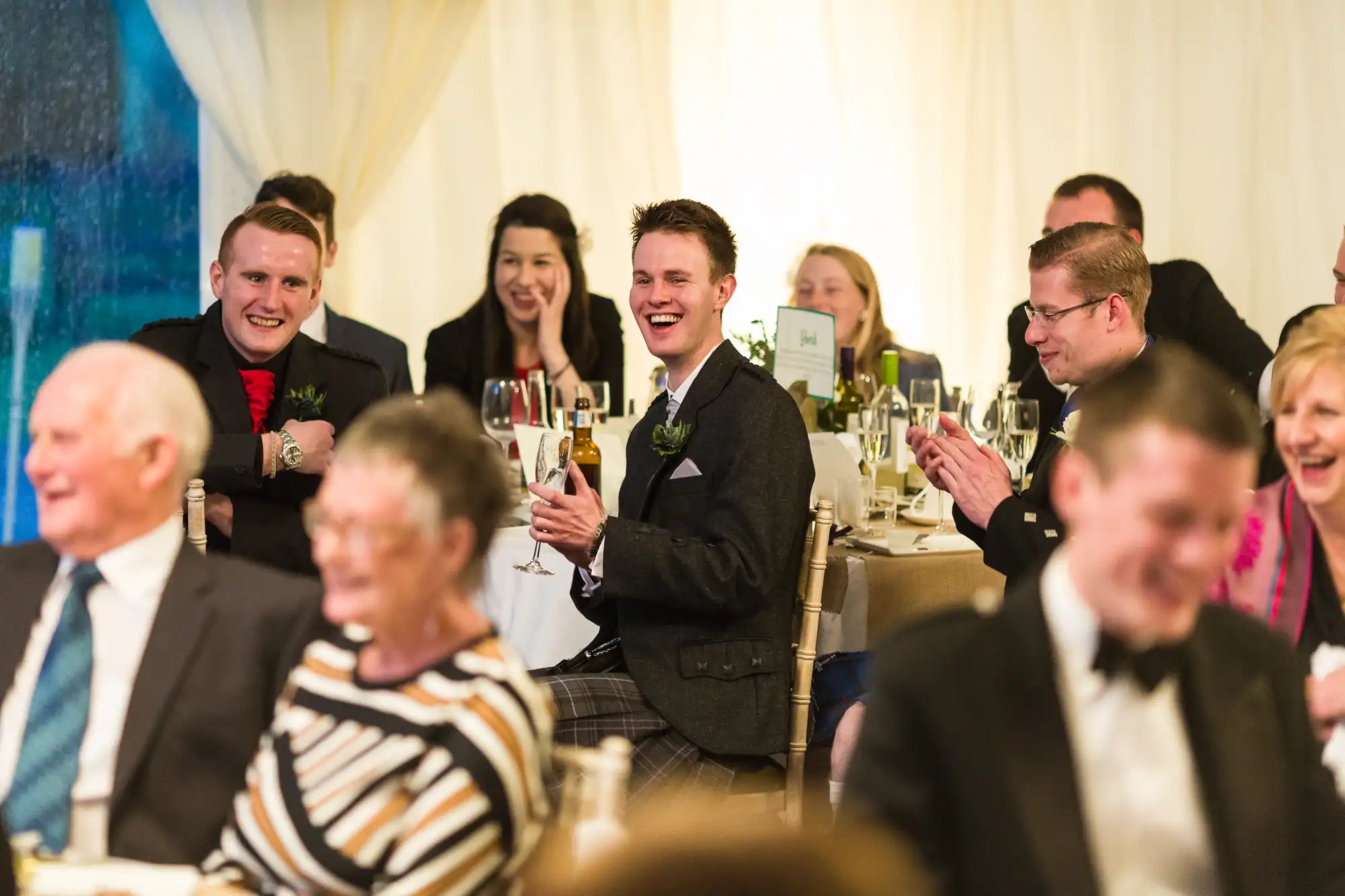 A group of people dressed formally sit at tables, smiling, laughing, and clapping at an indoor event. The main focus is on a young man in a dark suit in the centre, holding a glass and laughing. The background shows festive lighting and blurred guests.