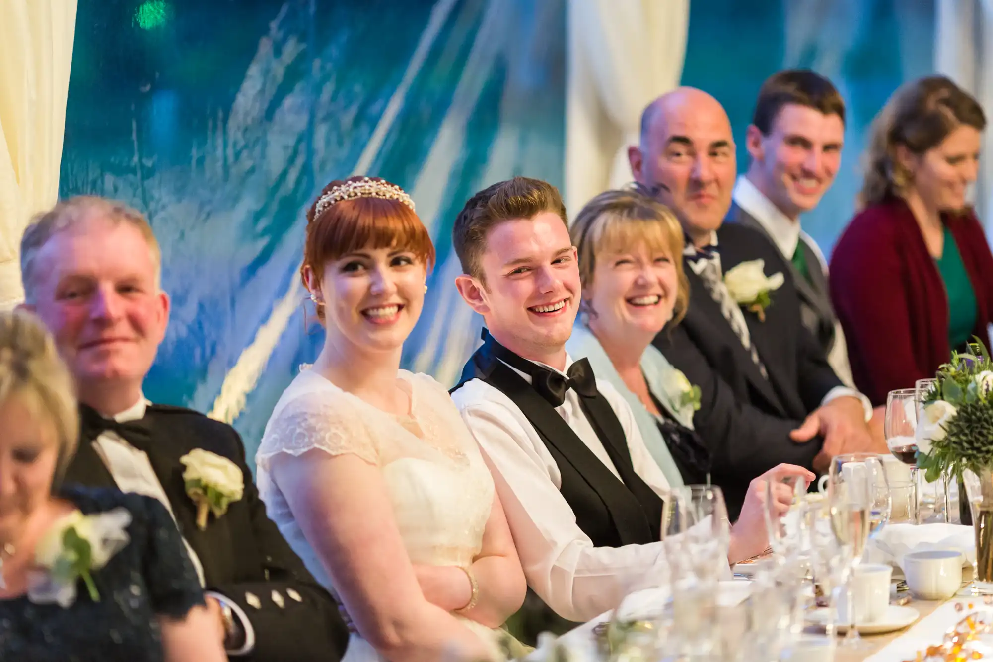 A smiling bride and groom sit together at a long, decorated table with guests at a wedding reception. Everyone is dressed formally, laughing and looking towards the camera, with glasses and floral arrangements in front of them.