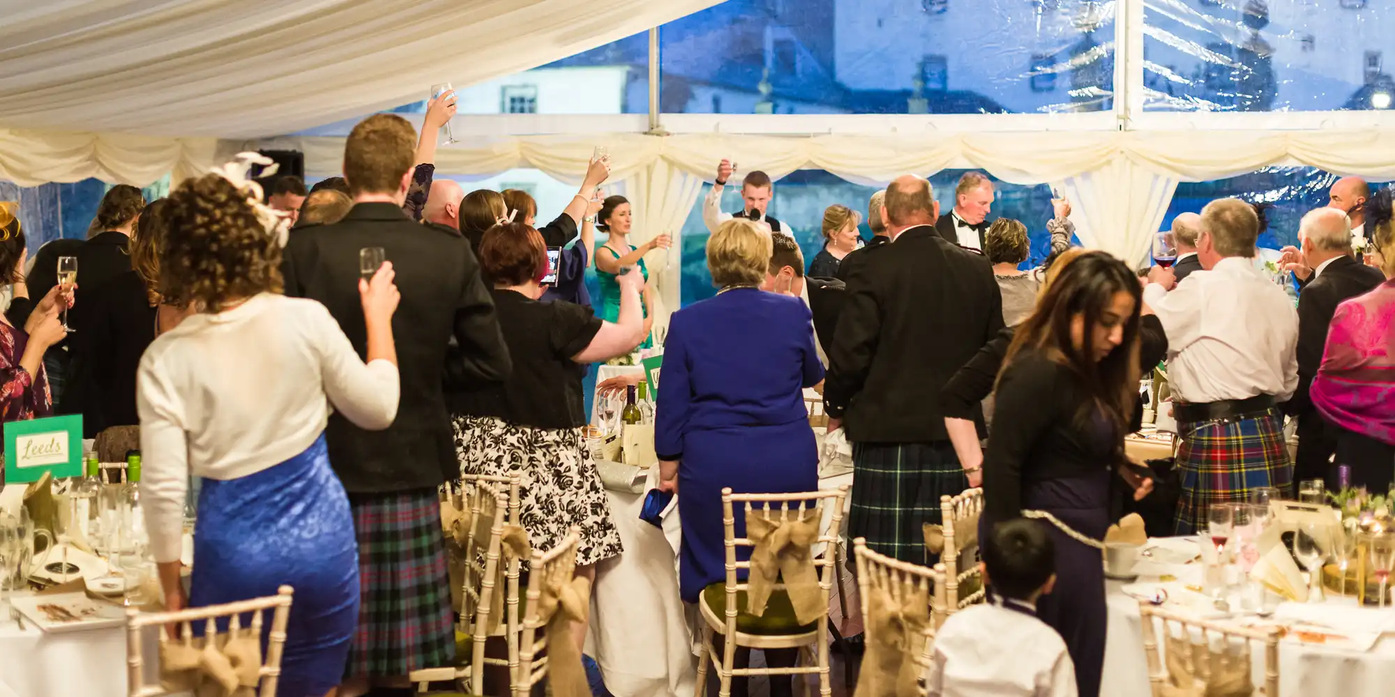 People in formal attire, many men in kilts, stand and celebrate at a decorated indoor event with round tables and white chairs. Some guests raise their arms or glasses. The atmosphere is lively under a white marquee with windows showing an evening view.