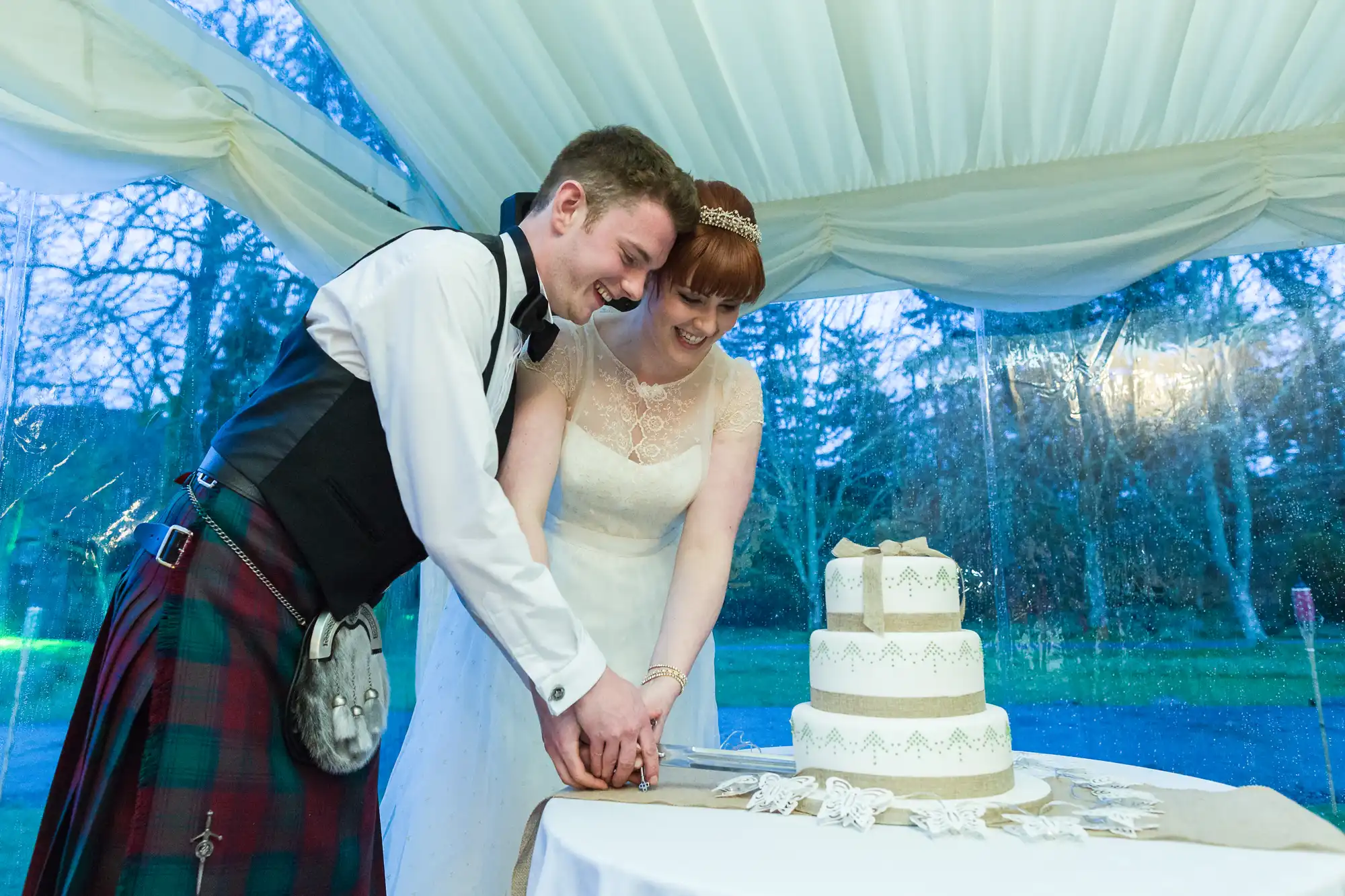 A bride in a white dress and a groom in a kilt and waistcoat smile as they cut a three-tiered white wedding cake together under a white marquee. The cake is decorated with lace-like icing, hessian, and small bows. Trees are visible outside through clear marquee panels.