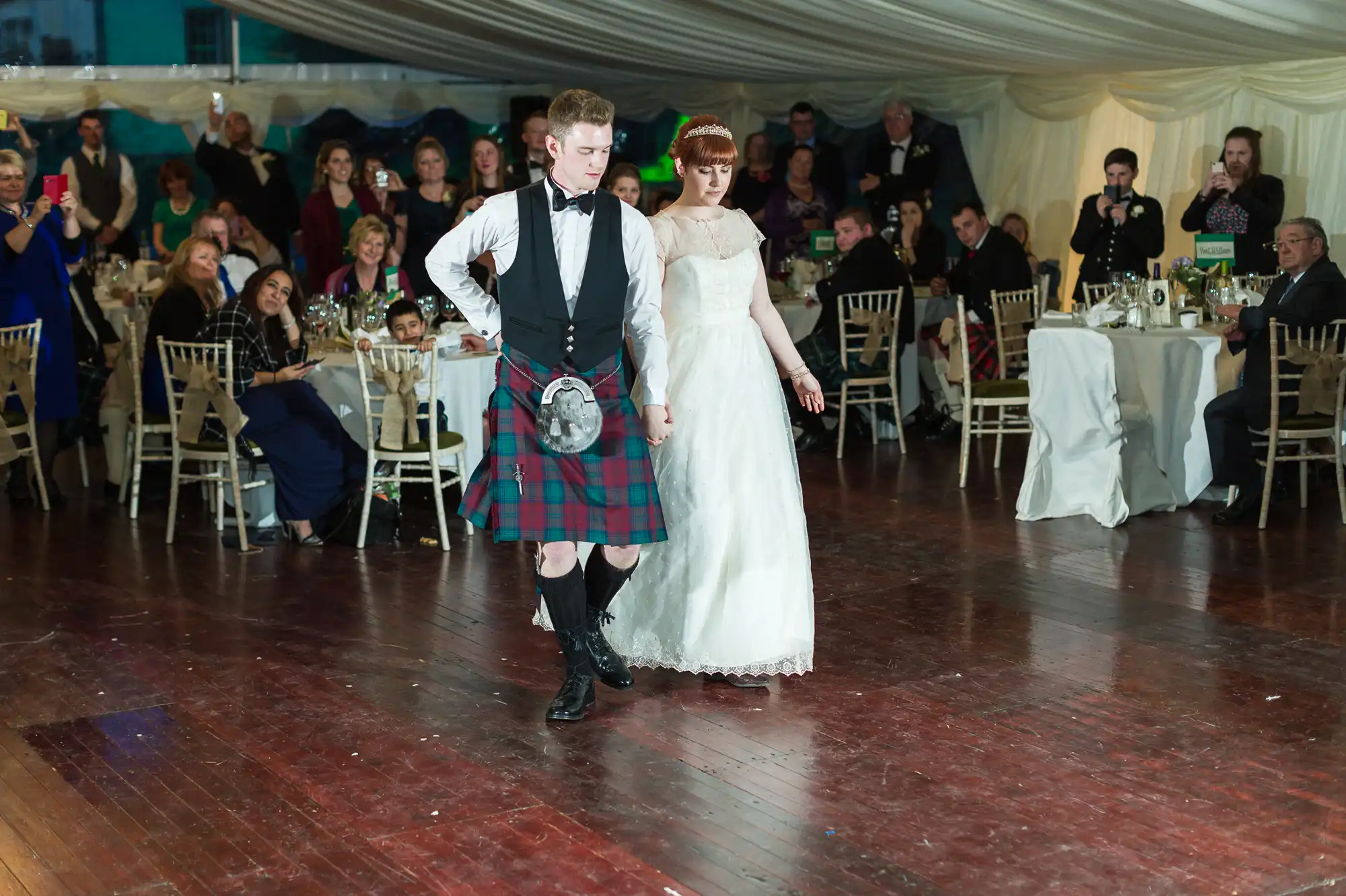 A bride in a white dress and a groom in a tartan kilt and bow tie dance together on a wooden floor in a marquee wedding venue, surrounded by seated and standing guests watching and taking photos.
