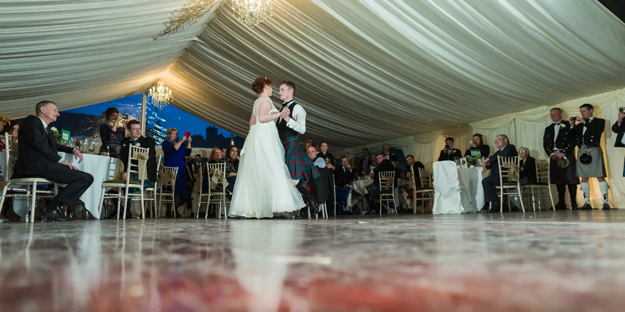 A bride and groom dance together in the centre of a large, elegant marquee with a draped ceiling and chandeliers, as seated and standing guests watch and take photos. Some guests wear formal attire, including kilts. The floor is shiny and reflective.