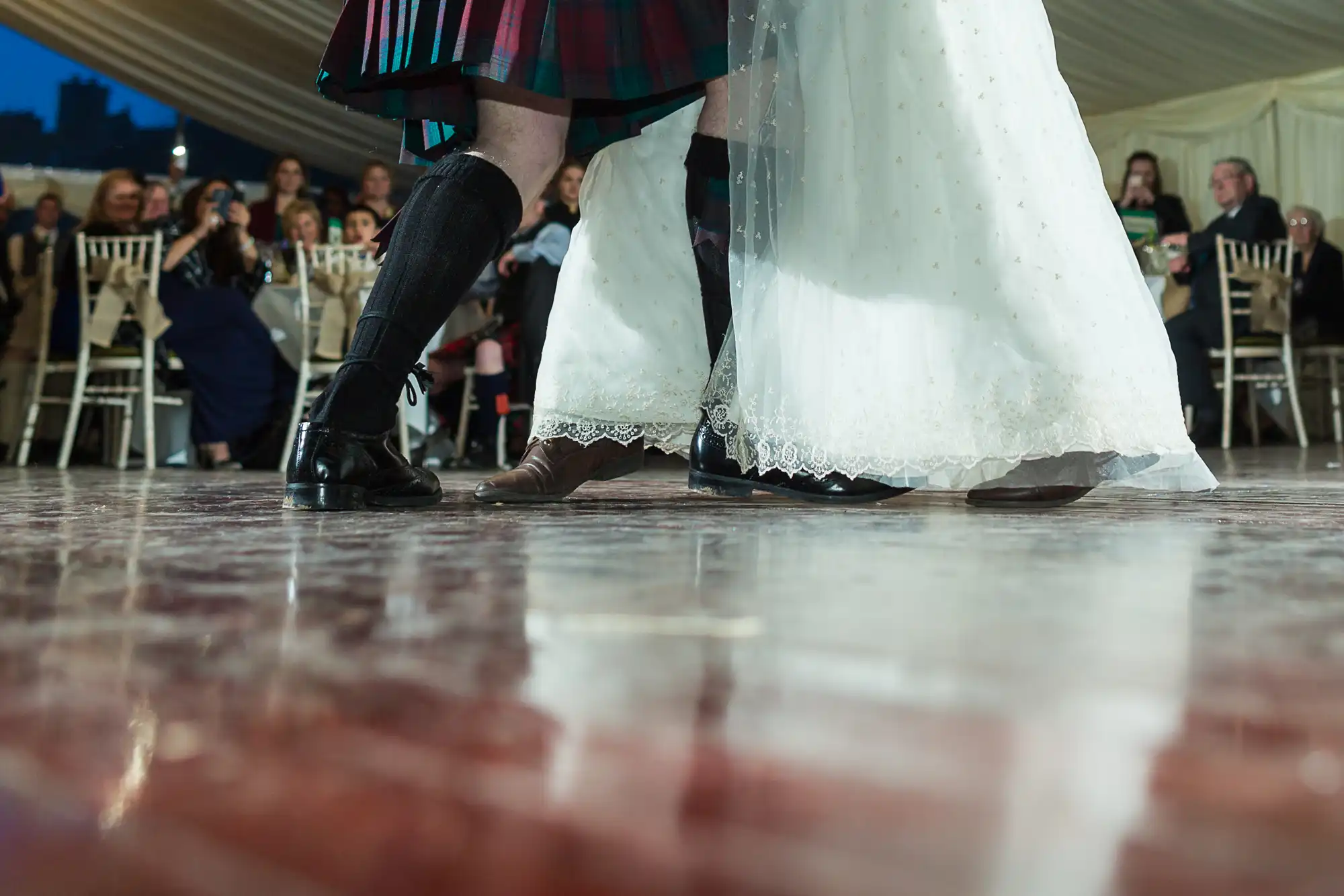 A close-up of a couple dancing at a wedding; one person wears a white dress and the other a tartan kilt. Their feet are mid-step on a shiny wooden floor, with seated guests and a white-draped marquee in the softly focused background.