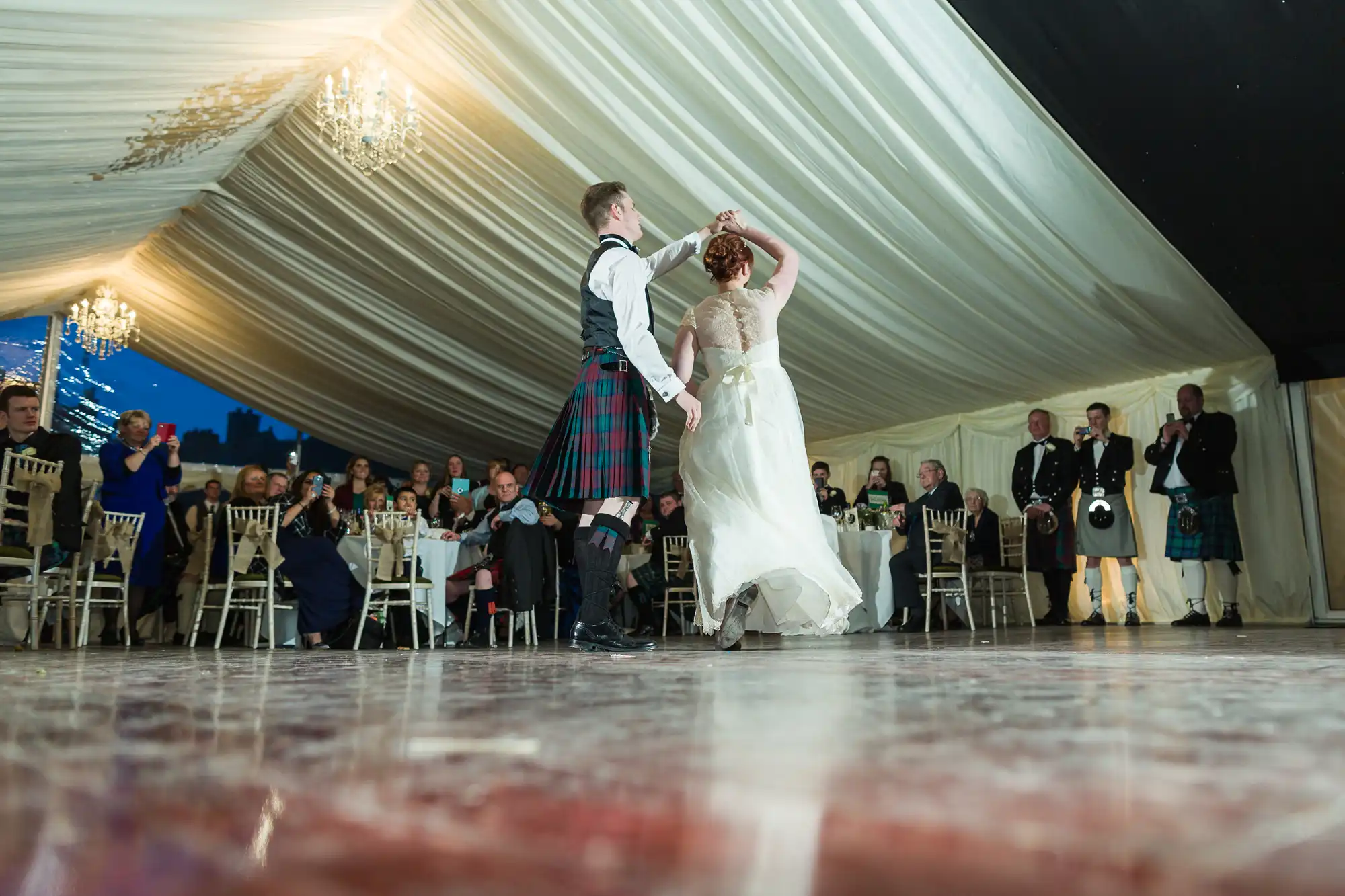 A couple dances on a polished floor in a decorated marquee; the man wears a kilt and the woman a white dress. Guests seated at round tables watch, and some people in kilts stand near the wall under chandeliers and draped white ceilings.