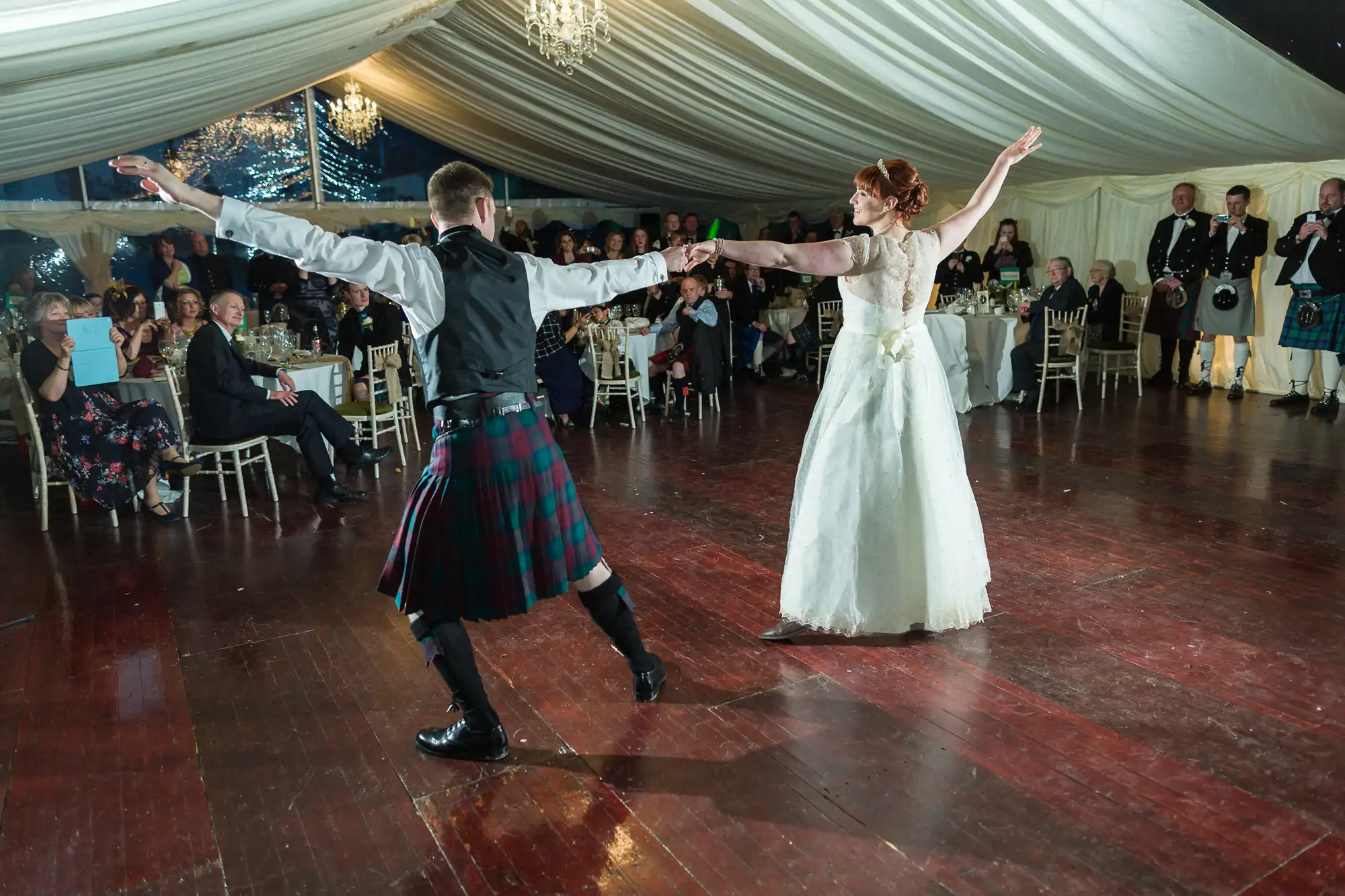 A bride in a white gown and a groom in a kilt dance with arms outstretched on a polished wooden floor in a large, elegant marquee. Guests watch from round tables, and men in kilts stand near the wall. Chandeliers hang from the marquee ceiling.
