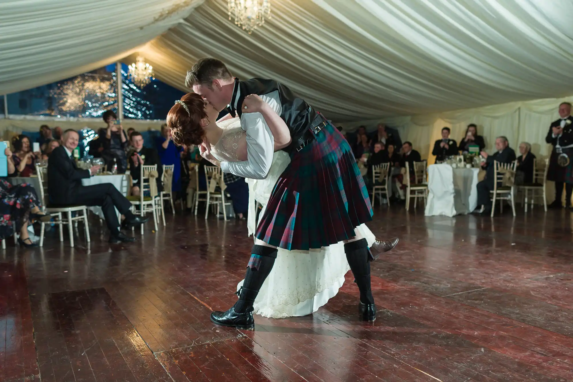 A couple dances in a tented venue; the man in a tartan kilt dips the woman in a white wedding dress. Guests seated at round tables watch, and chandeliers hang from the draped ceiling, creating an elegant, celebratory atmosphere.