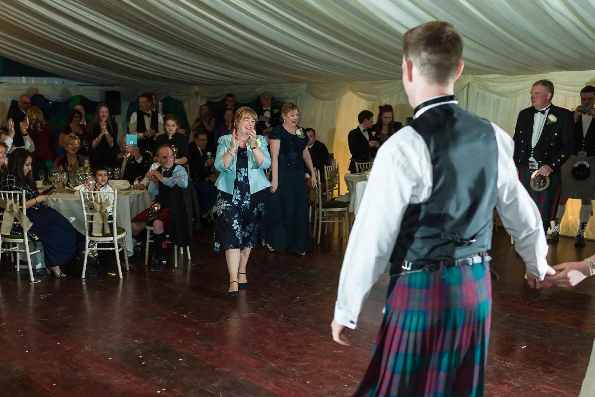 A smiling woman in a blue dress walks across a dancefloor toward a man in a tartan kilt and waistcoat, who holds someone’s hand. Guests seated at tables and standing in a decorated marquee watch the scene, some clapping and taking photos.