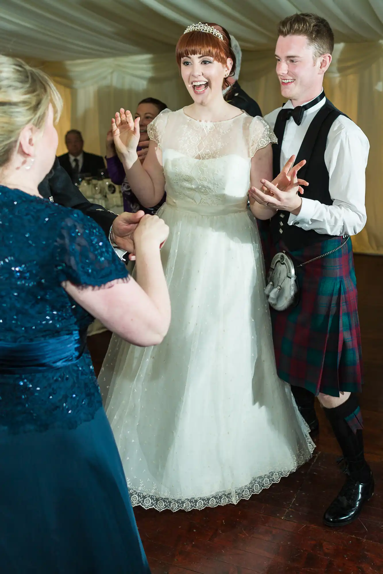 A smiling bride in a white dress and a groom in a kilt dance together at a wedding reception. Another woman in a navy blue dress faces them, raising her hands. Other seated guests are visible in the background under a white marquee.