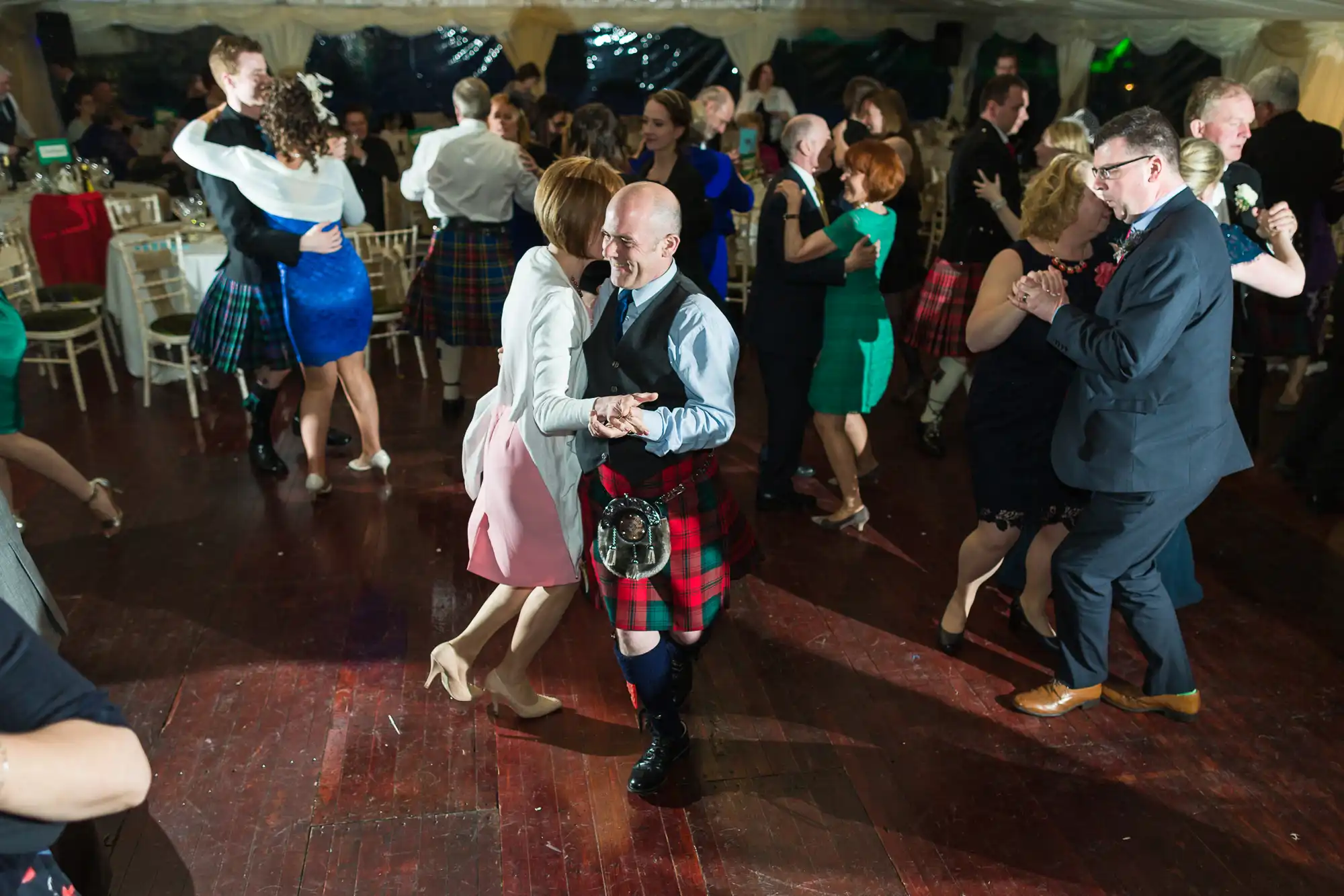 Couples dance on a wooden floor at a lively event; many men wear tartan kilts and formal jackets, while women wear dresses. The room is crowded, with people smiling and engaged, suggesting a festive Scottish celebration.