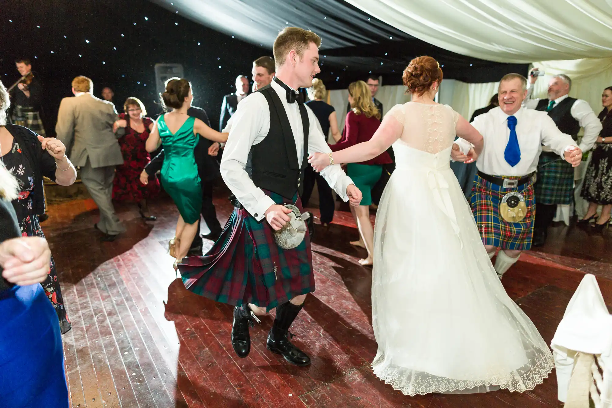 People in formal attire, including men in kilts and a woman in a white wedding dress, dance joyfully on a wooden floor under a draped marquee with twinkling lights. They hold hands and smile, celebrating in a lively, festive atmosphere.
