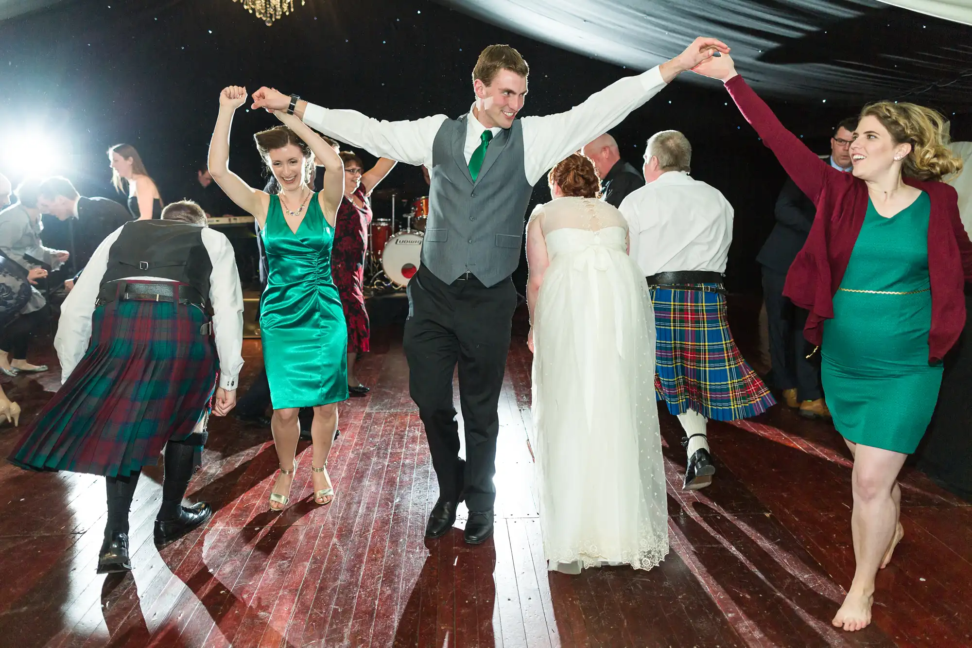 People in colourful formal attire, including kilts, suits, dresses, and a wedding dress, dance energetically on a polished wooden floor under a draped ceiling, holding hands and smiling. A bright light shines from the background.