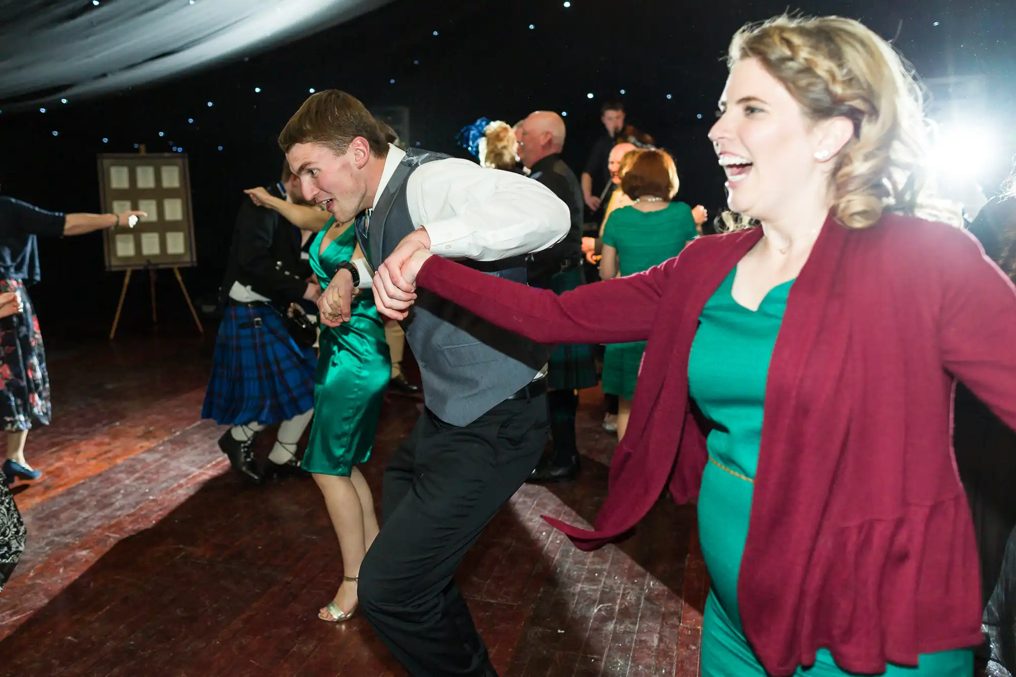 A group of people hold hands and dance energetically at a festive indoor event. The foreground shows a smiling woman in a green dress and maroon cardigan, and a man in a waistcoat. The background is filled with other dancers and bright event lighting.