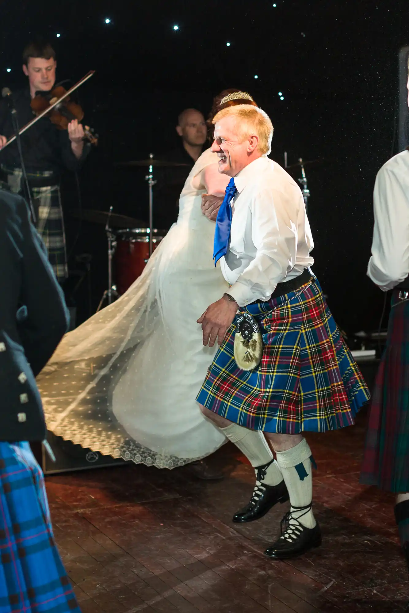 An older man wearing a tartan kilt, white shirt, blue tie, and sporran dances joyfully at an event. A bride in a white dress twirls nearby. Musicians play in the background, and other dancers in kilts are partially visible. The scene is lively and festive.