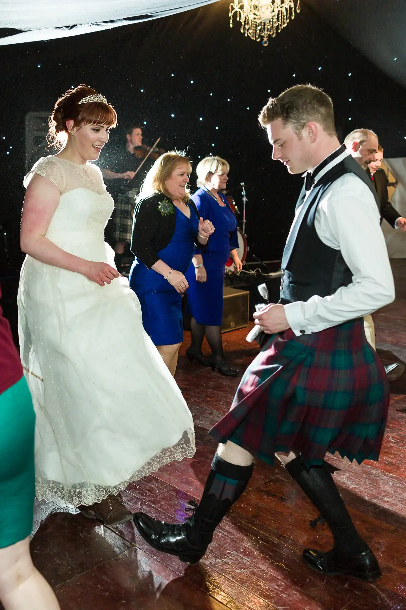 A bride in a white gown and a man in a tartan kilt dance energetically at a wedding reception. Other guests, dressed in formal attire, watch and smile in the background. The floor is wooden and a chandelier hangs above. The atmosphere is festive and joyful.