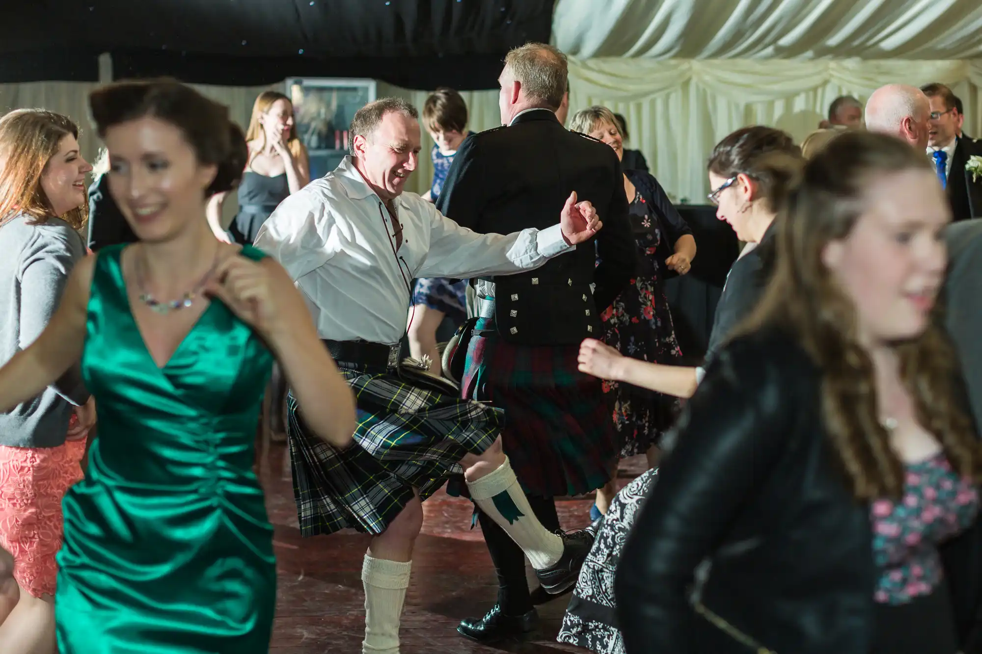 A lively group of people dance together at a celebration. Several men wear kilts and formal jackets, while women are in dresses. One man in the centre lifts his leg mid-step, smiling. The atmosphere is festive, with blurred motion showing energetic dancing.