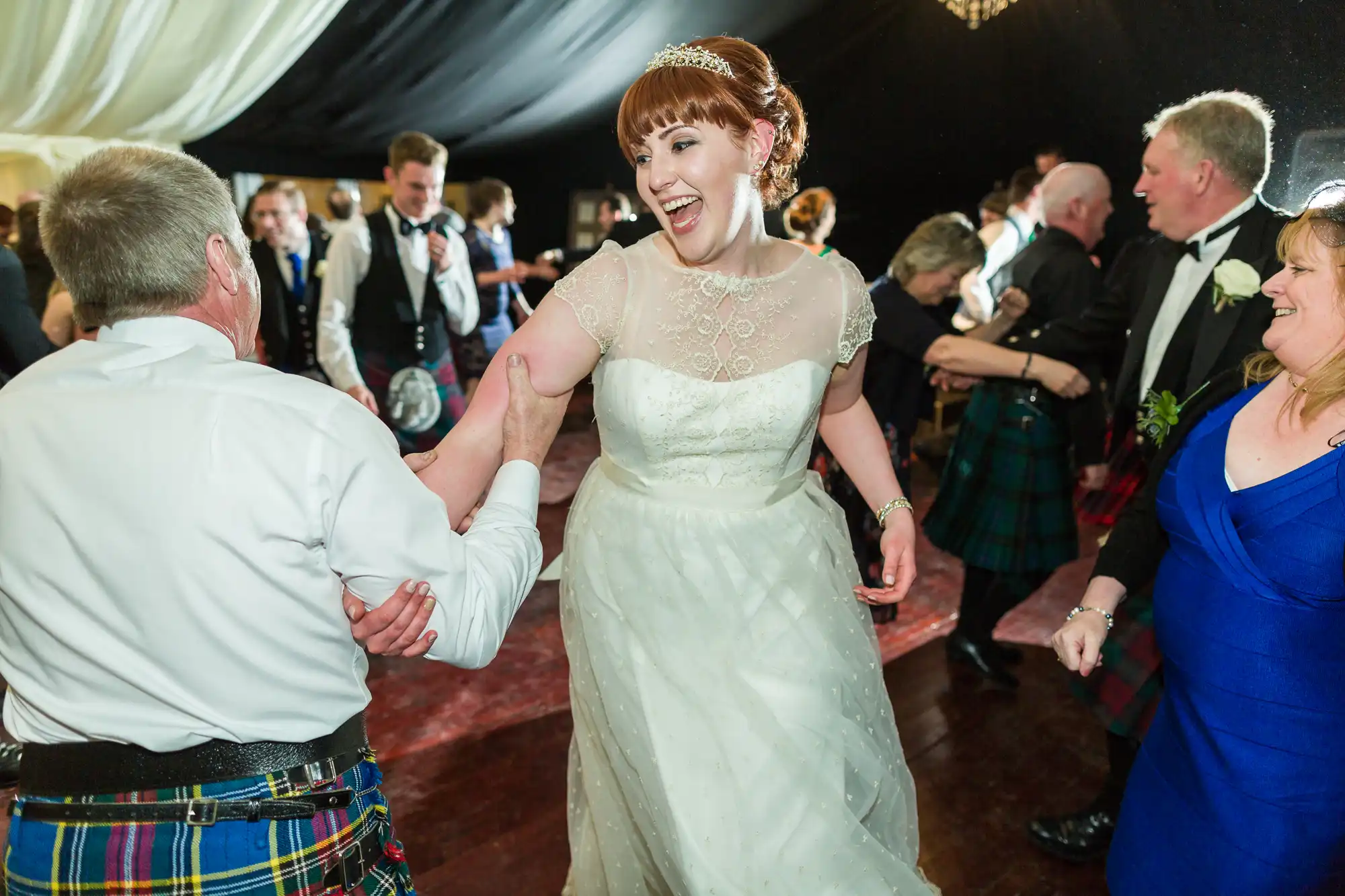 A joyful bride in a white dress and tiara laughs as she dances, holding hands with a man in a kilt. Wedding guests in formal attire, some wearing kilts, dance and celebrate around her under a marquee with draped fabric overhead.
