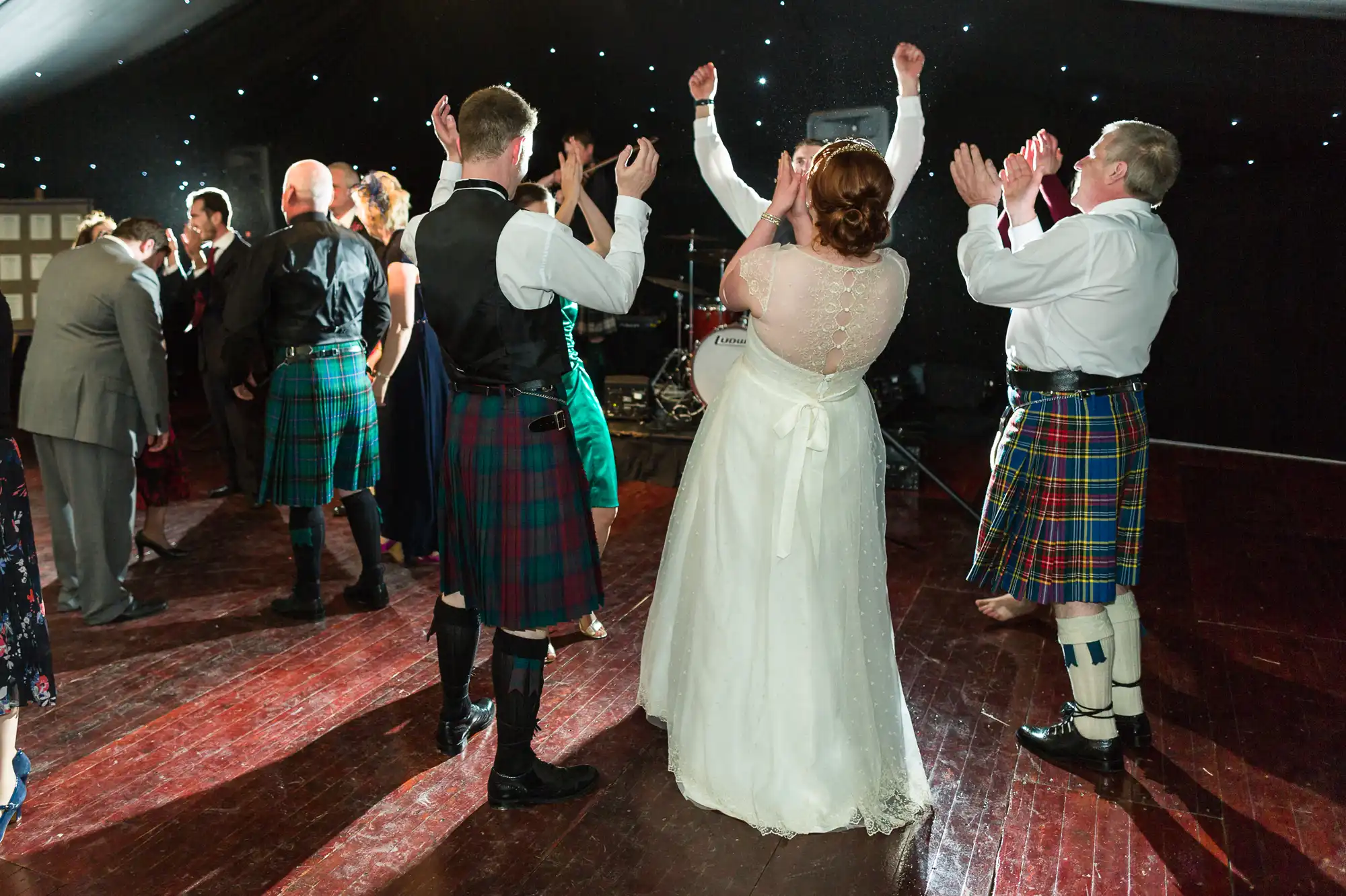 A bride in a white dress dances with guests, many wearing kilts, on a polished wooden floor. People clap and raise their hands. The background is decorated with fairy lights, creating a festive atmosphere.