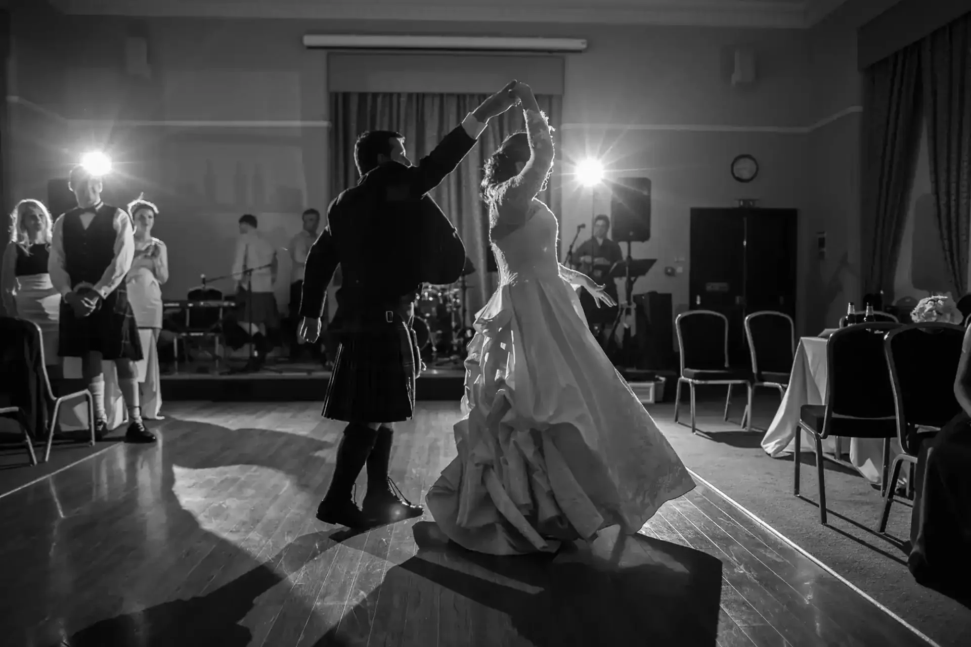 A bride and groom dance in the centre of a dimly lit ballroom at Upper and Lower College Halls at the University of St Andrews, illuminated by spotlights. The bride wears a flowing white gown and the groom wears a kilt. Guests stand and watch in the background. Empty chairs and round tables line the room's edge.