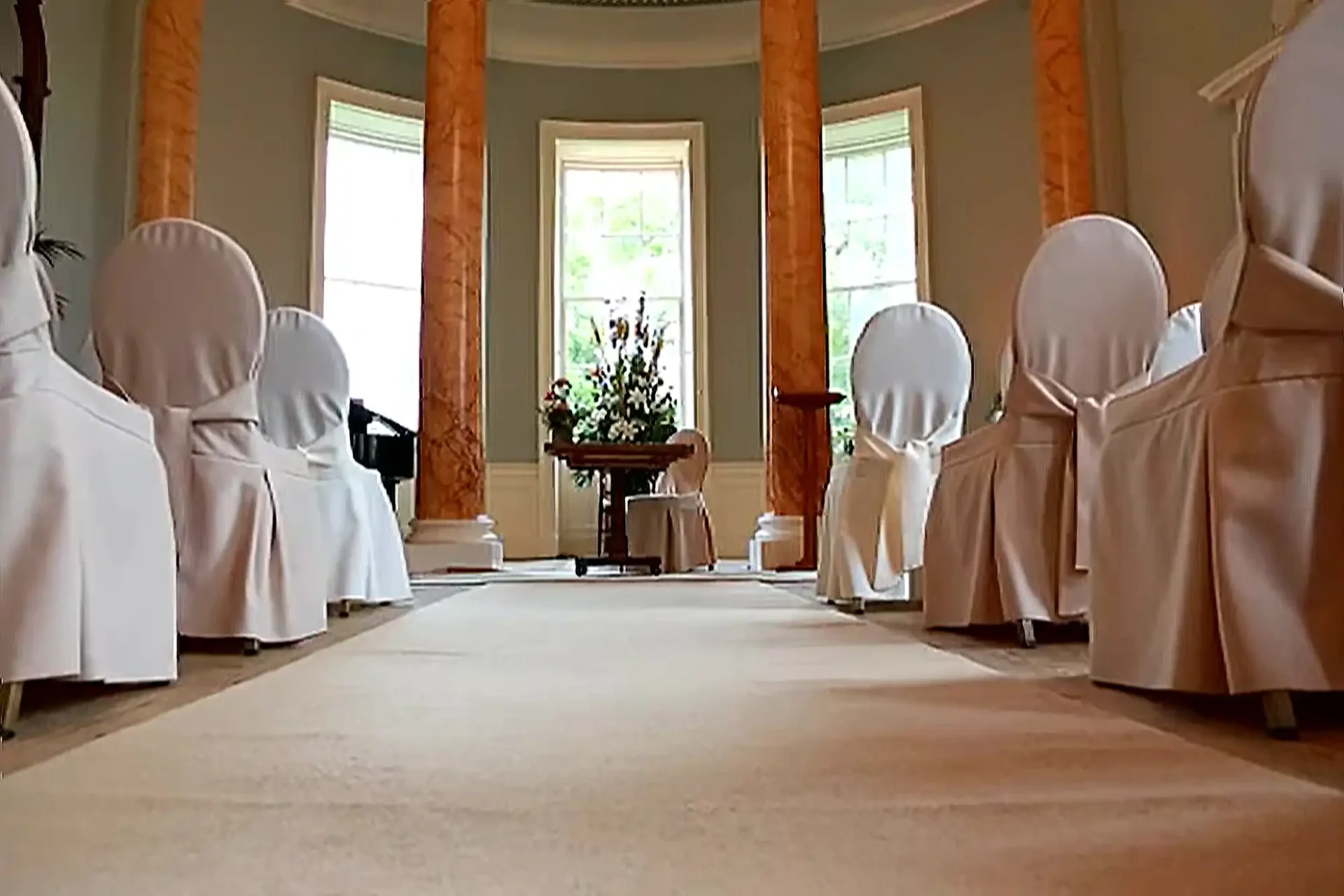 A formal room set for a wedding at Wedderburn Castle, featuring two rows of white-covered chairs facing a central table with a large floral arrangement. Three tall windows and marble columns frame the back wall, whilst soft natural light illuminates the elegant, carpeted space.
