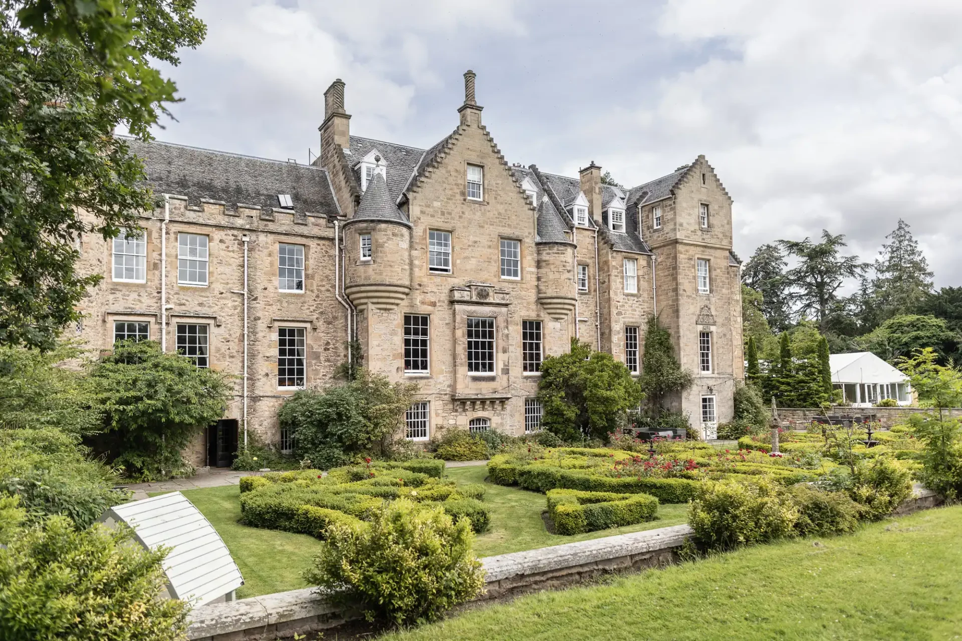 Historic Carberry Tower with gabled roofs and turrets, surrounded by manicured gardens and hedges, under a cloudy sky.