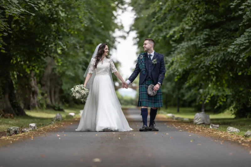 wedding photographer at Carberry Tower: A bride in a white gown and a groom in a kilt walk hand in hand down a tree-lined path.