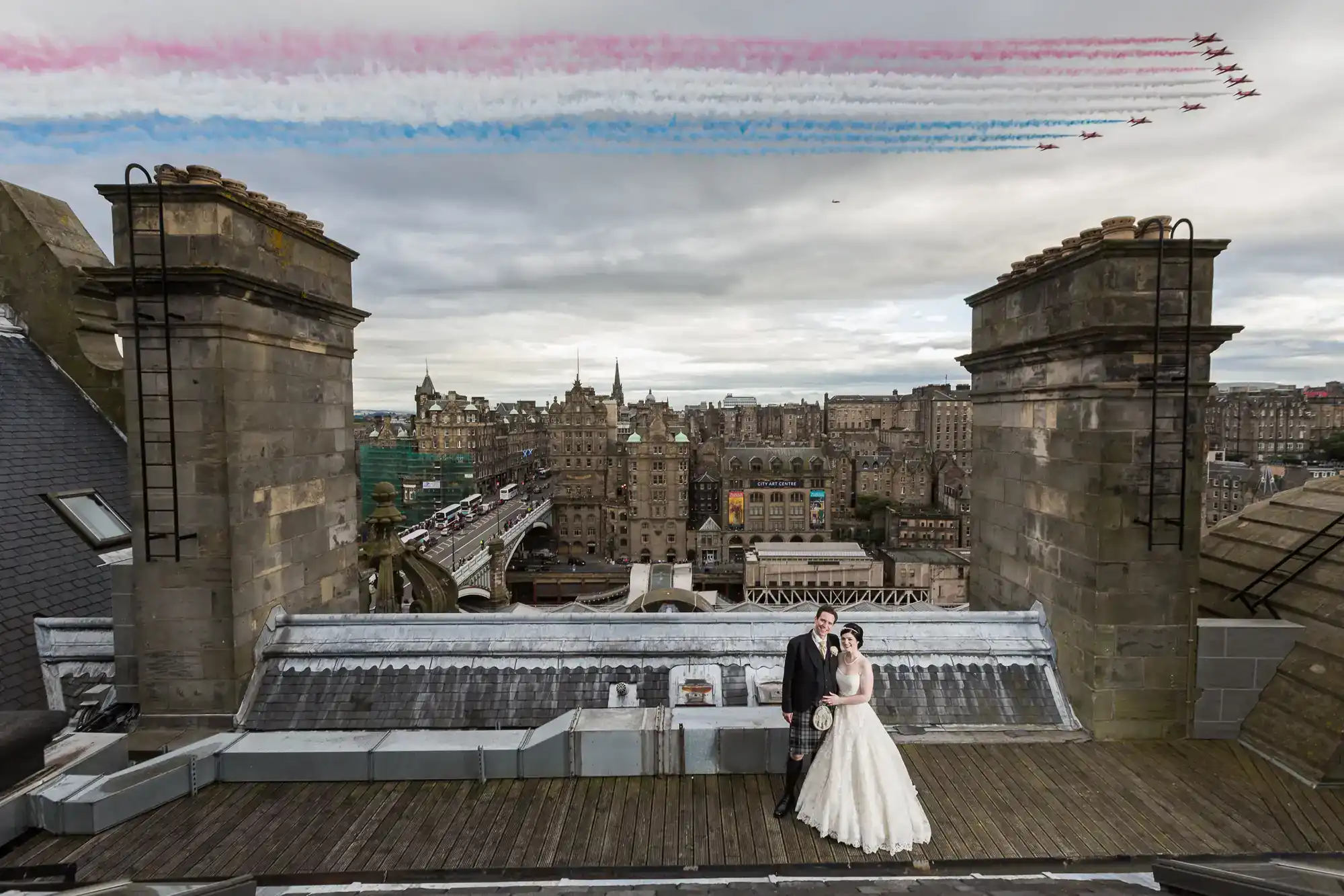 A bride and groom stand on a rooftop with a cityscape in the background, while the Red Arrows planes fly overhead leaving red, white, and blue smoke trails in the sky, at the Balmoral Hotel, Edinburgh.