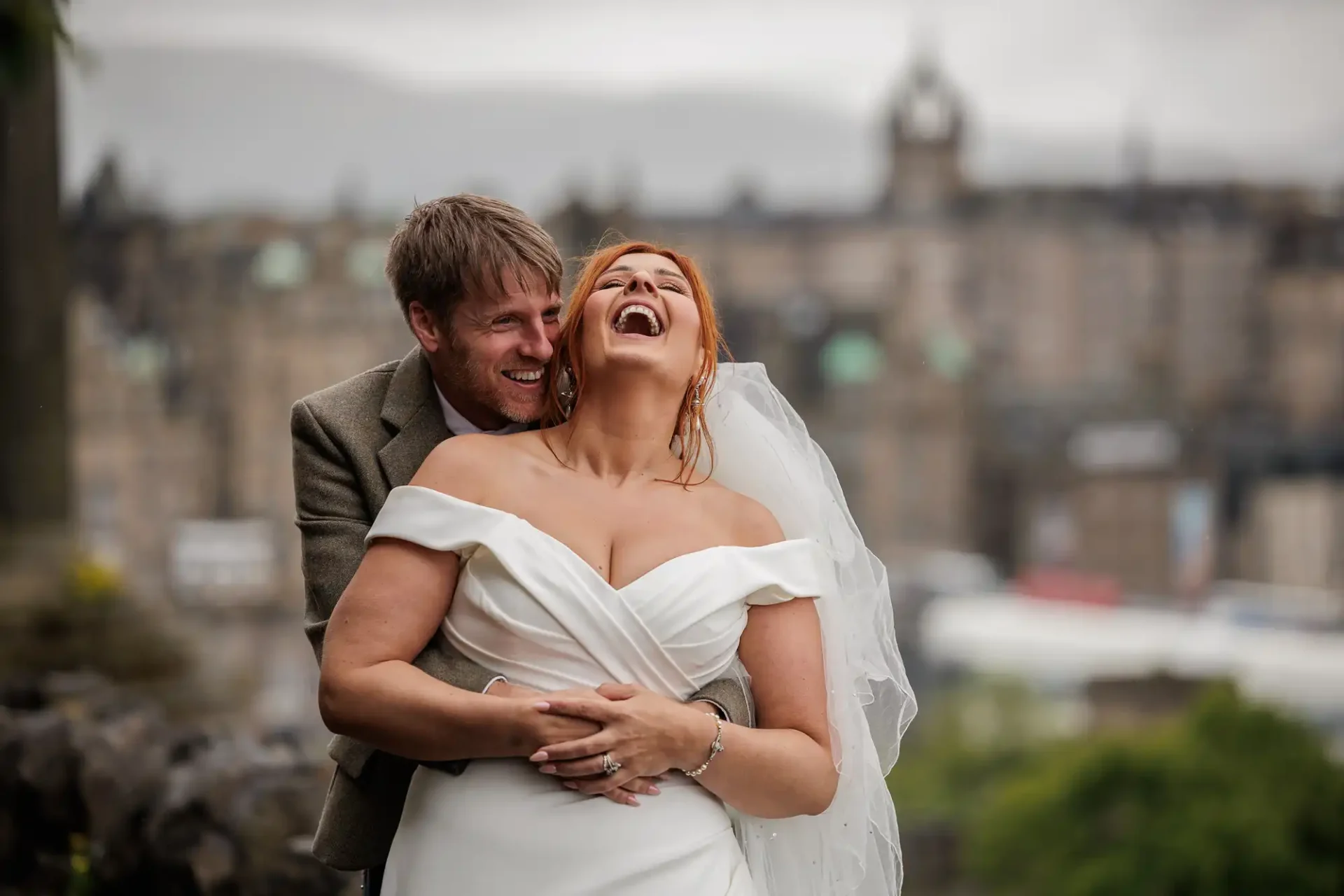 A bride in a white dress and veil laughs as a groom in a suit embraces her from behind outdoors, with blurred city buildings in the background at Calton Hill, Edinburgh.