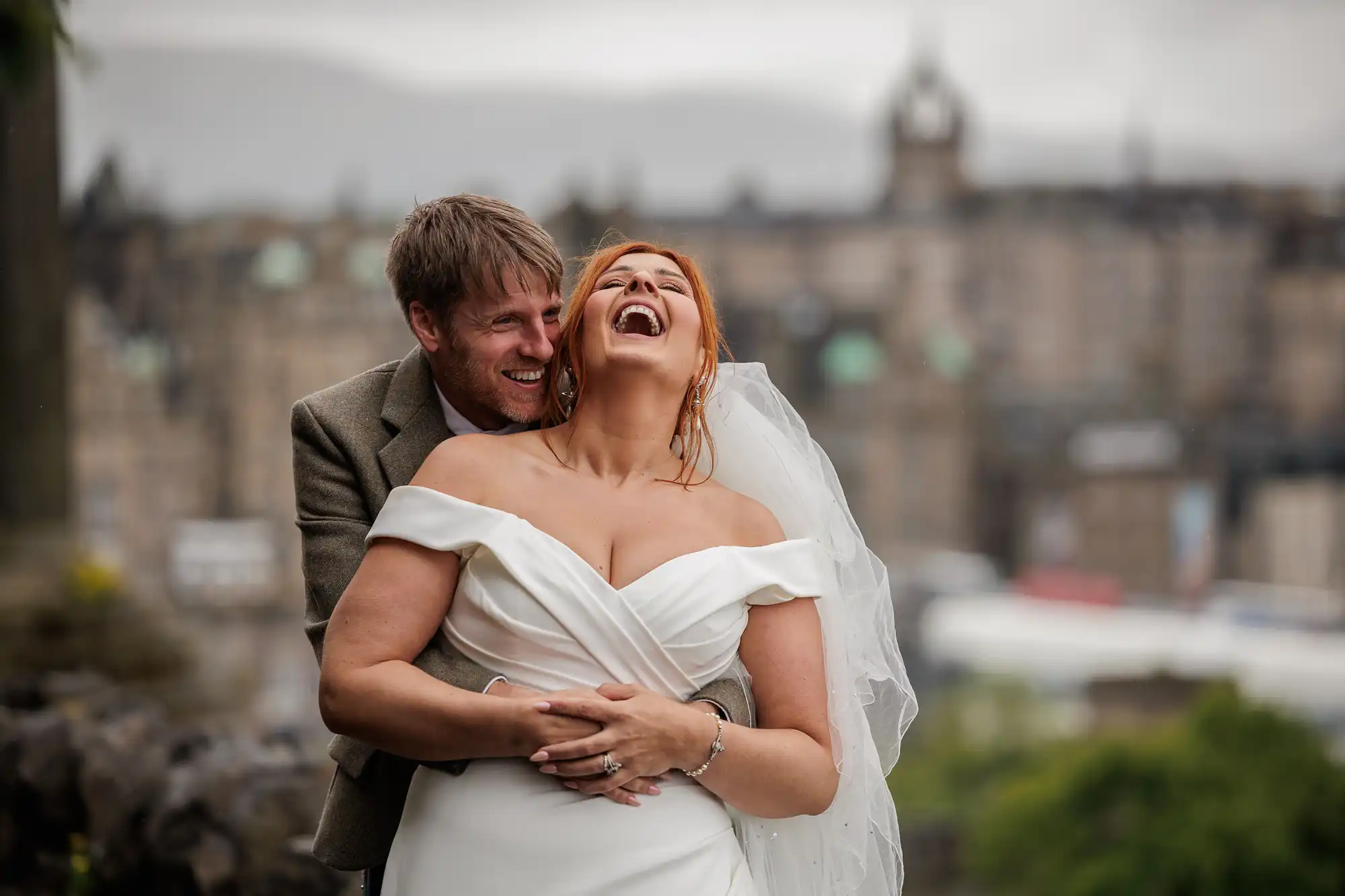 A bride in a white dress and veil laughs as a groom in a suit embraces her from behind outdoors, with blurred city buildings in the background at Calton Hill, Edinburgh.