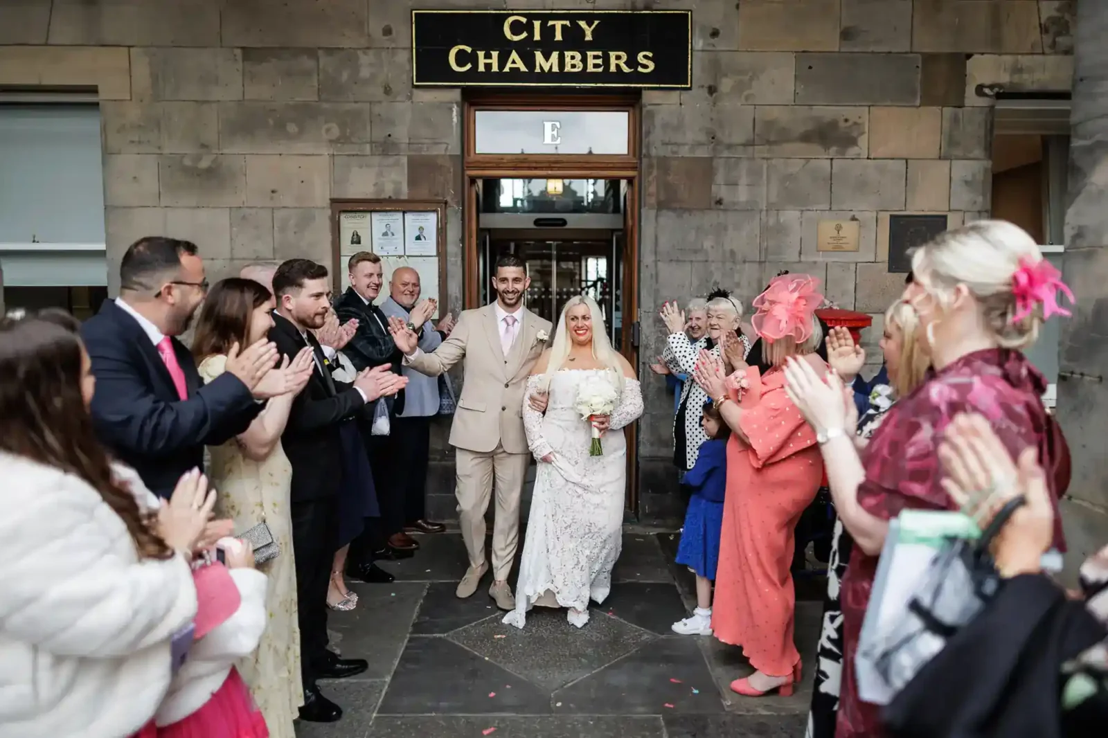 A newlywed couple in light-coloured attire stands smiling at the entrance of City Chambers, holding a bouquet. Family and friends line both sides, cheering and applauding in celebration. The building’s stone exterior and a City Chambers sign are visible above. A romantic and natural wedding photograph of a couple outside the Edinburgh City Chambers on the High Street, captured by Love Wedding Photos And Film.