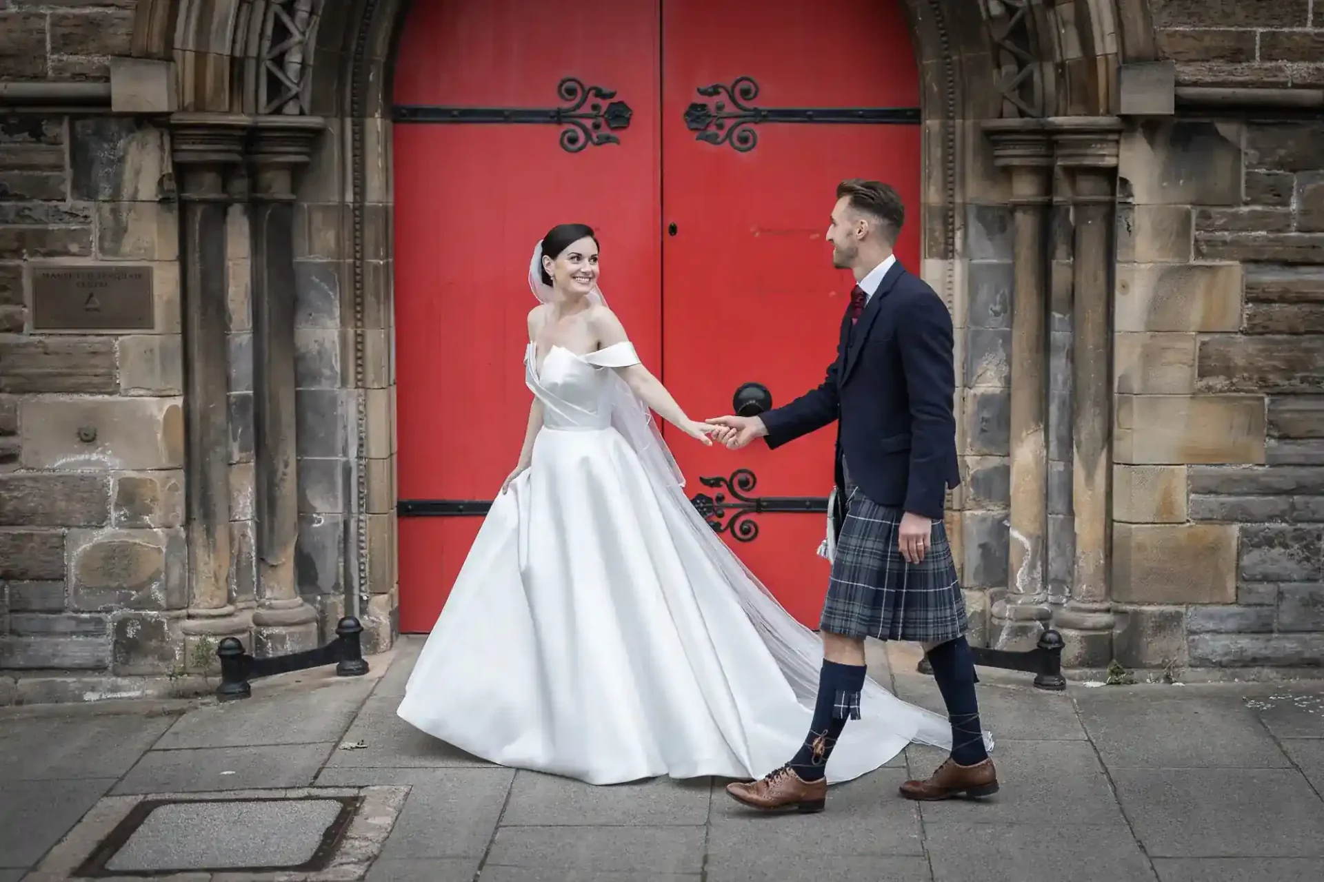 A bride in a white gown and a groom in a kilt hold hands and smile outside a stone building with large red doors at Mansfield Traquair Edinburgh.