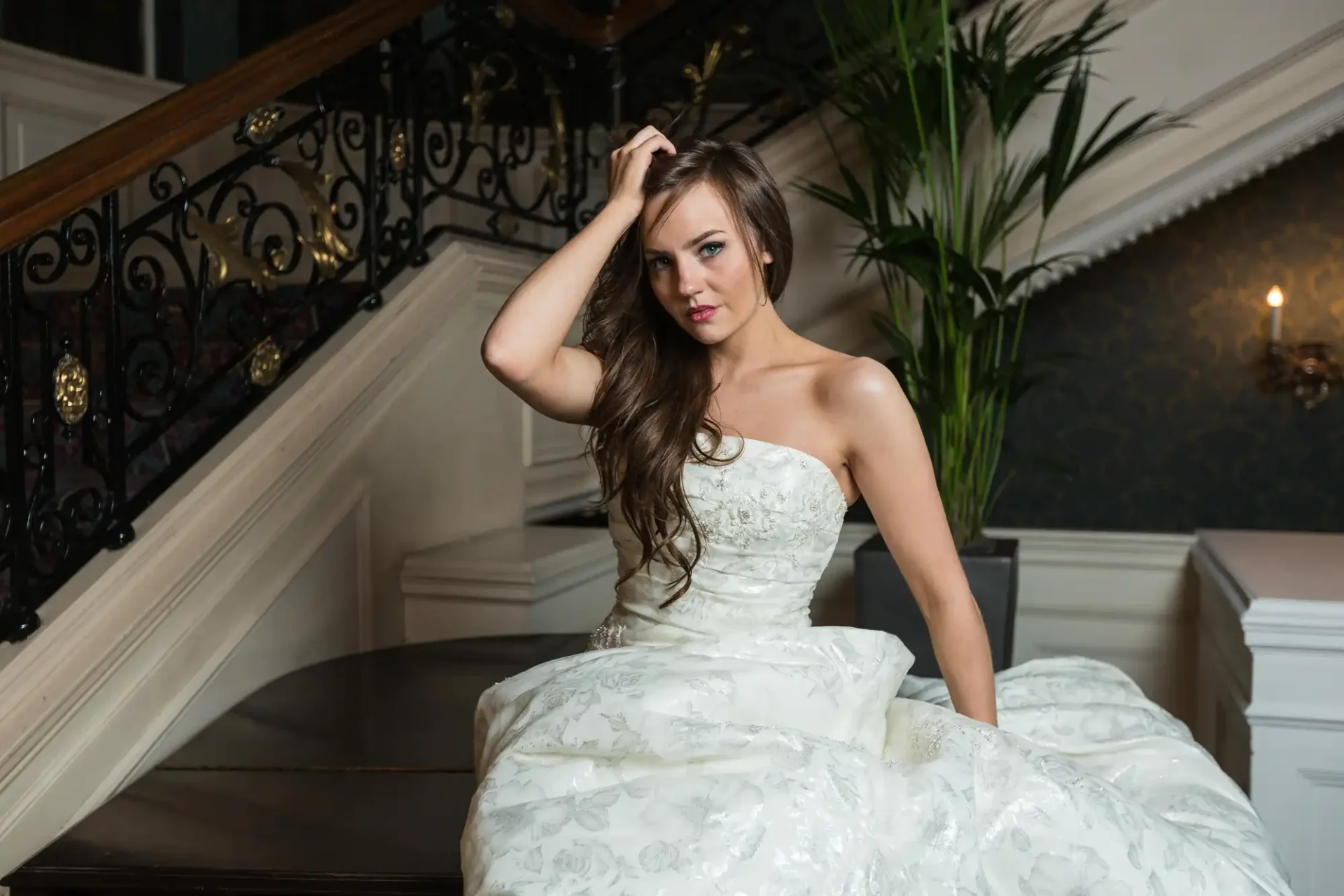 A woman in a strapless white wedding dress sits on a staircase landing, posing with one hand in her hair at the Balmoral Hotel, Edinburgh.