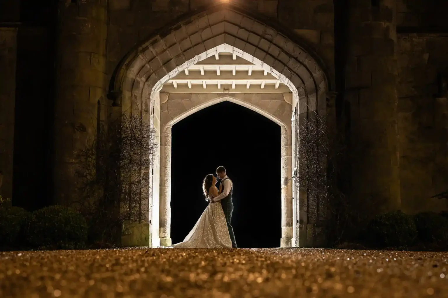 A bride and groom stand embraced under a large stone archway at night, illuminated by lights from behind at Dundas Castle, Edinburgh.