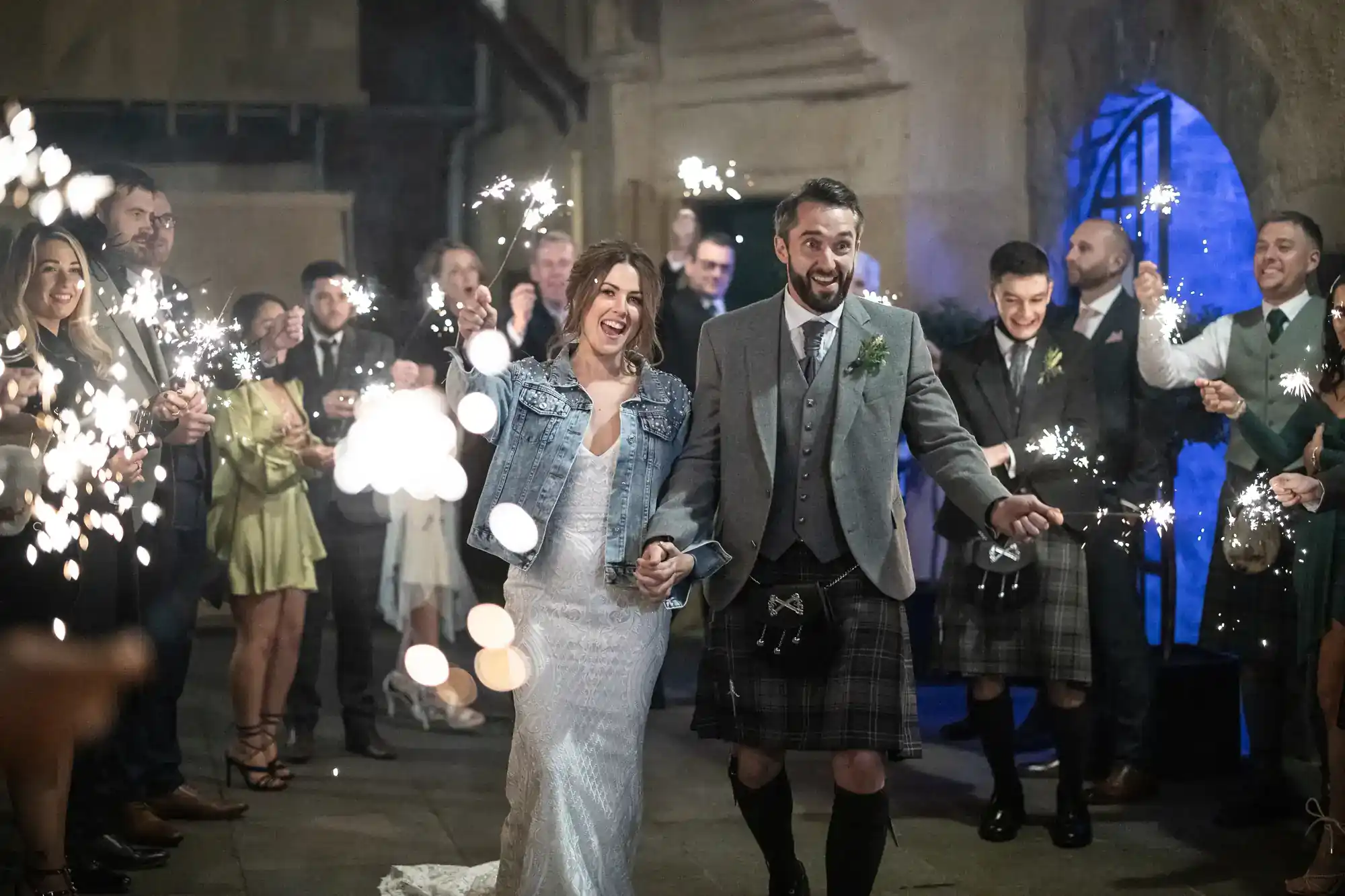 A bride and groom walk together smiling among guests holding sparklers at night; the groom wears a kilt and the bride wears a denim jacket over her dress at Riddles Court, Edinburgh.