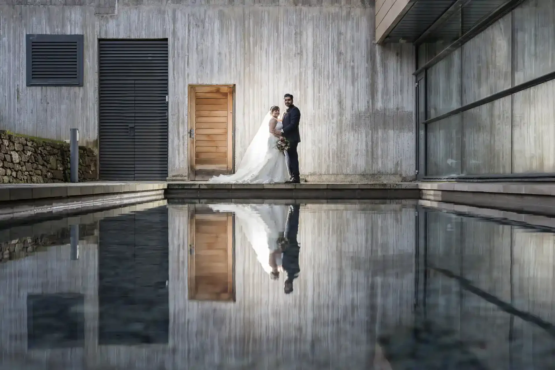 A bride and groom stand together in front of a modern, concrete building, with their reflection visible in a pool of water below at Norton House Hotel, Edinburgh.