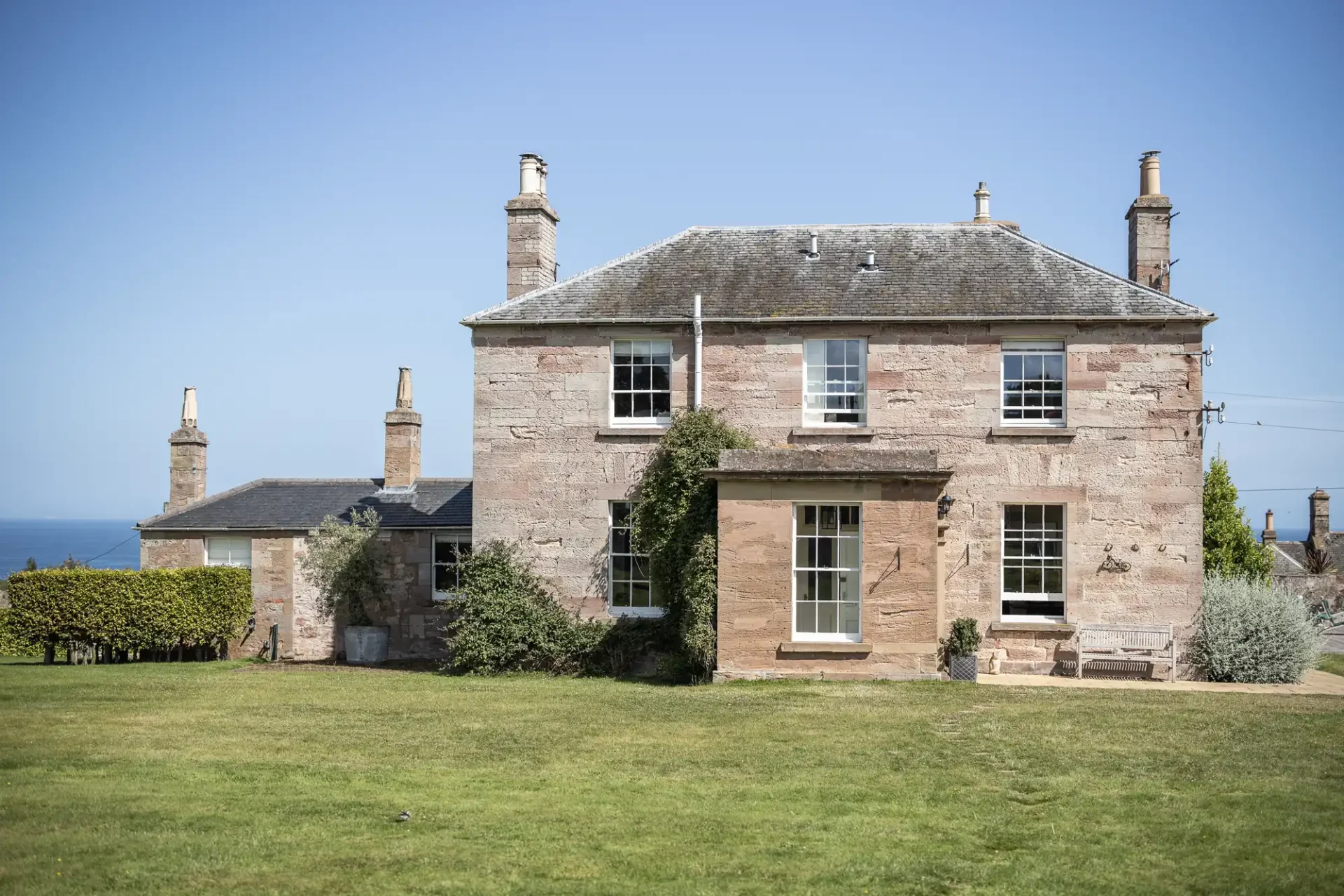 Two-story stone house with chimneys, surrounded by a grassy lawn, under a clear blue sky.