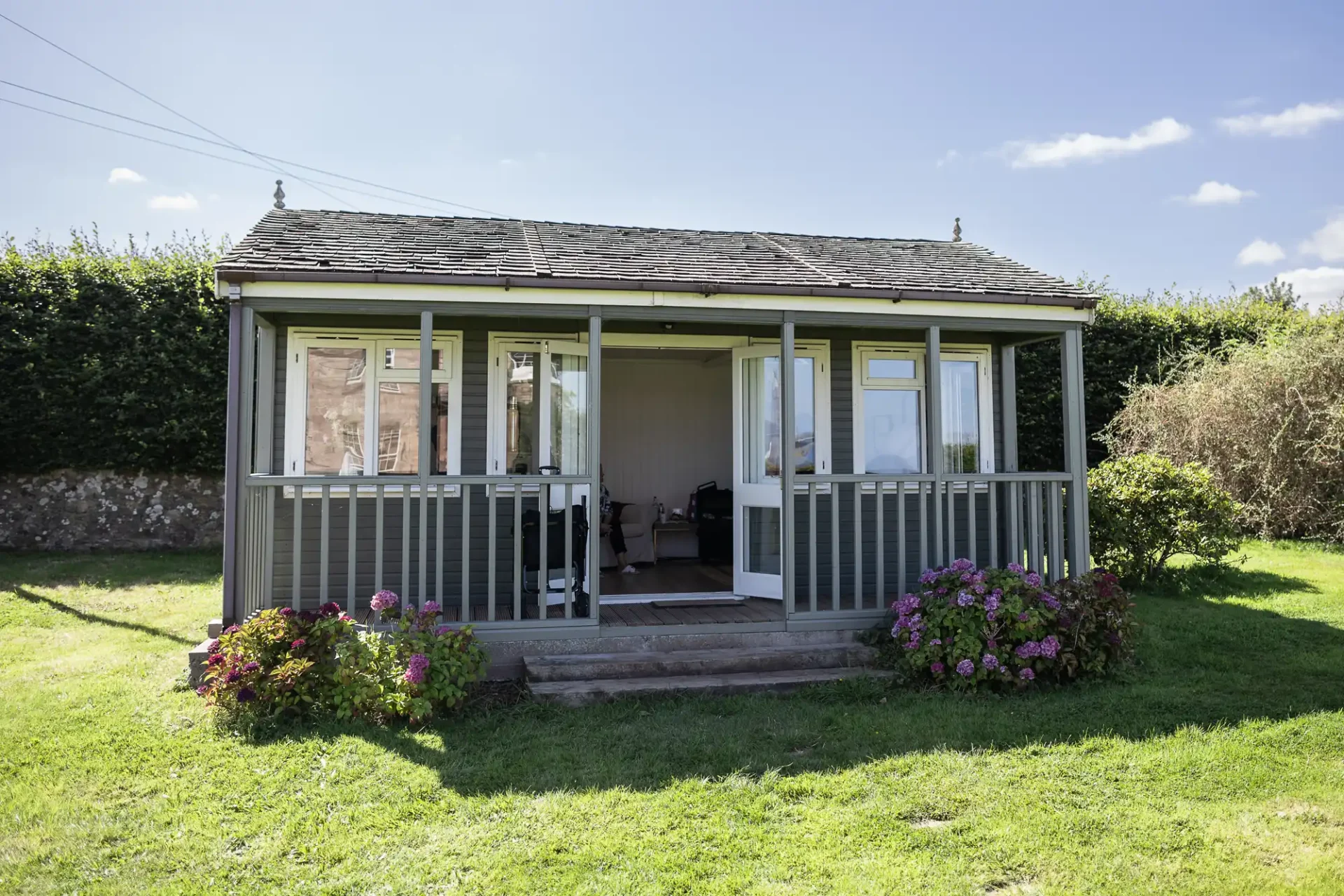 Small wooden cabin with large windows and a front porch, surrounded by grass and flowering bushes, under a clear blue sky.