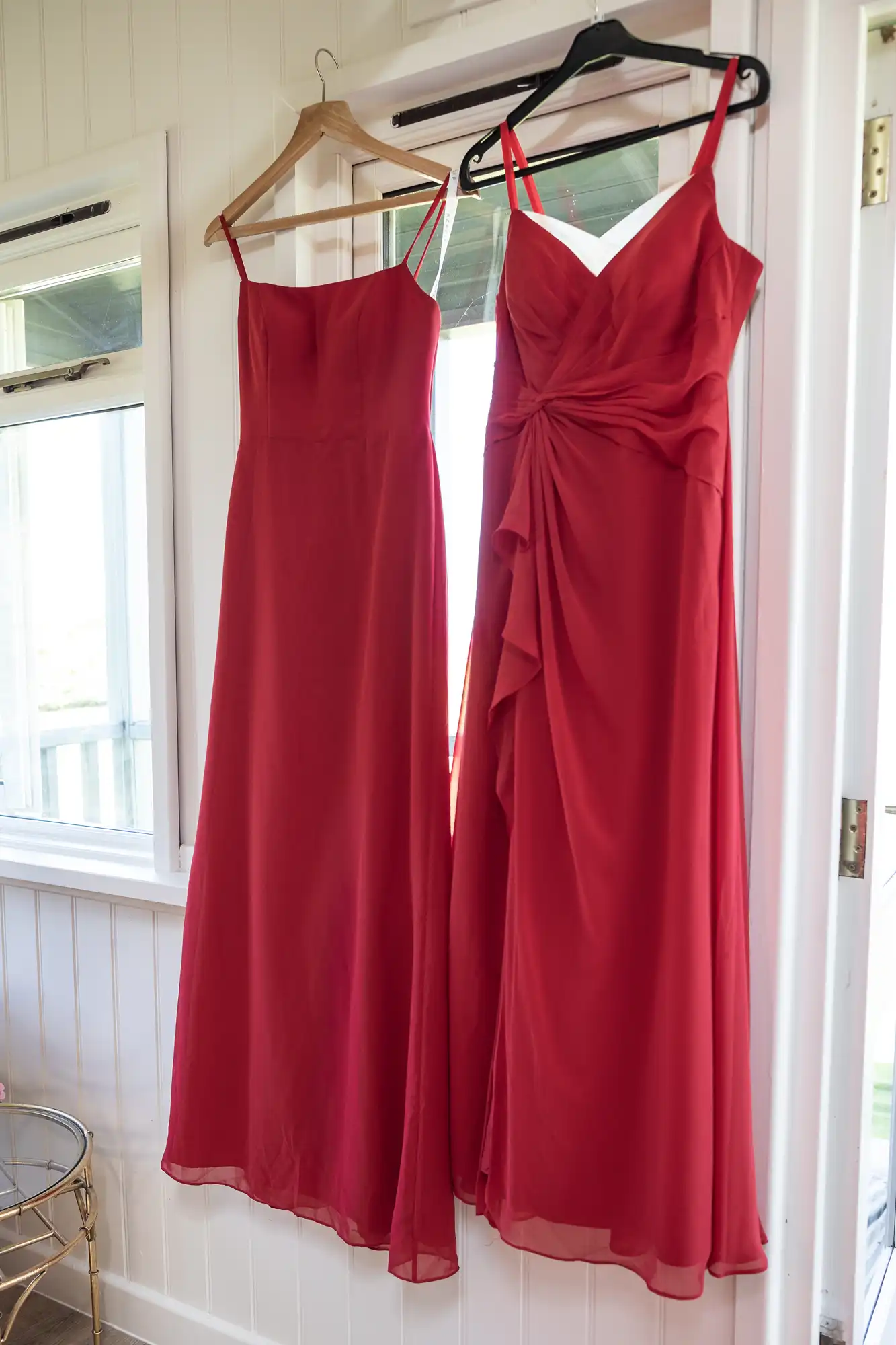 Two red dresses on hangers, displayed against a white wall with windows. The left dress is plain, while the right dress features a ruched design.